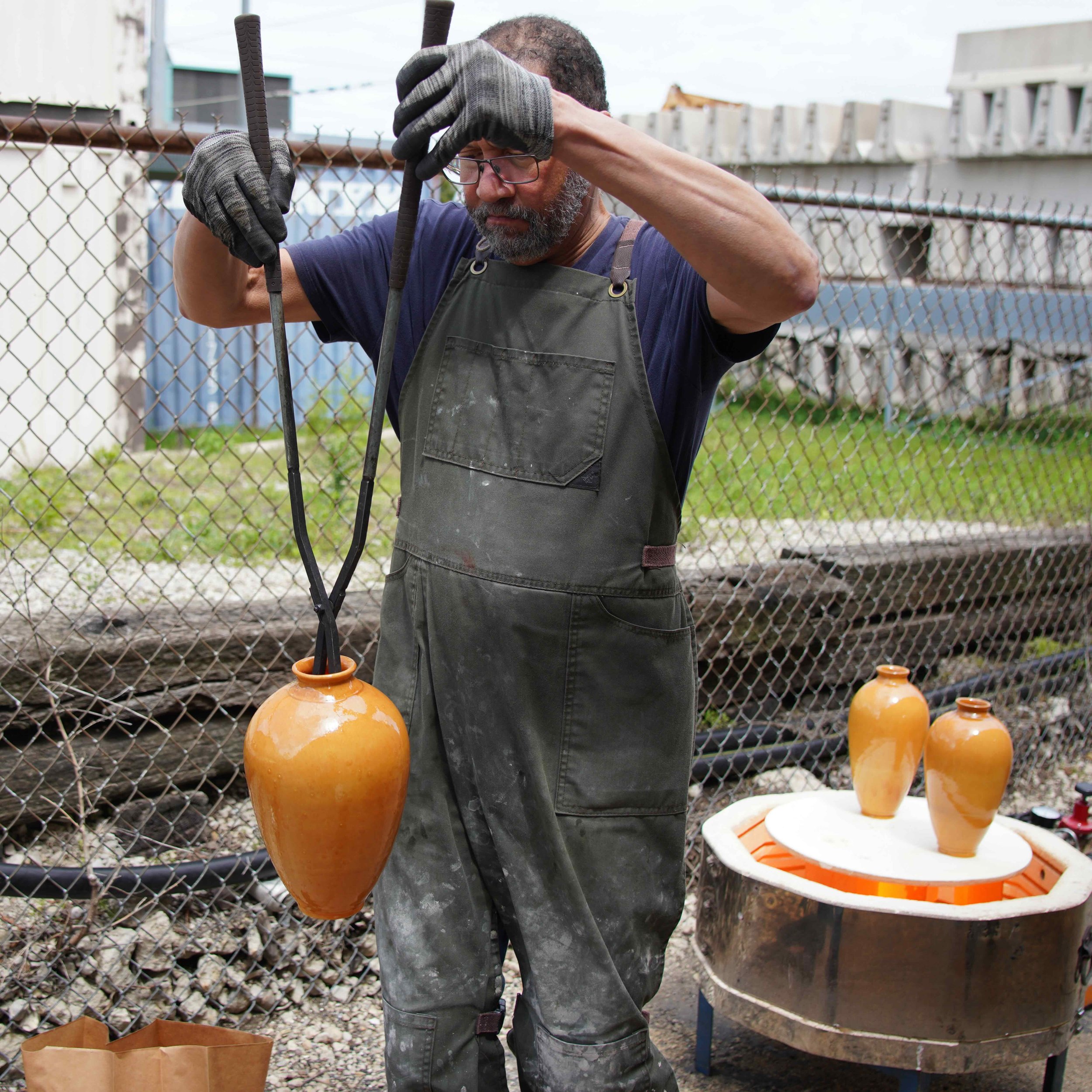 A man with a beard wearing glasses, a blue shirt, and an apron stands outdoors, with long tongs taking a glowing hot vessel from the kiln with several glowing hot ceramic vases behind him.