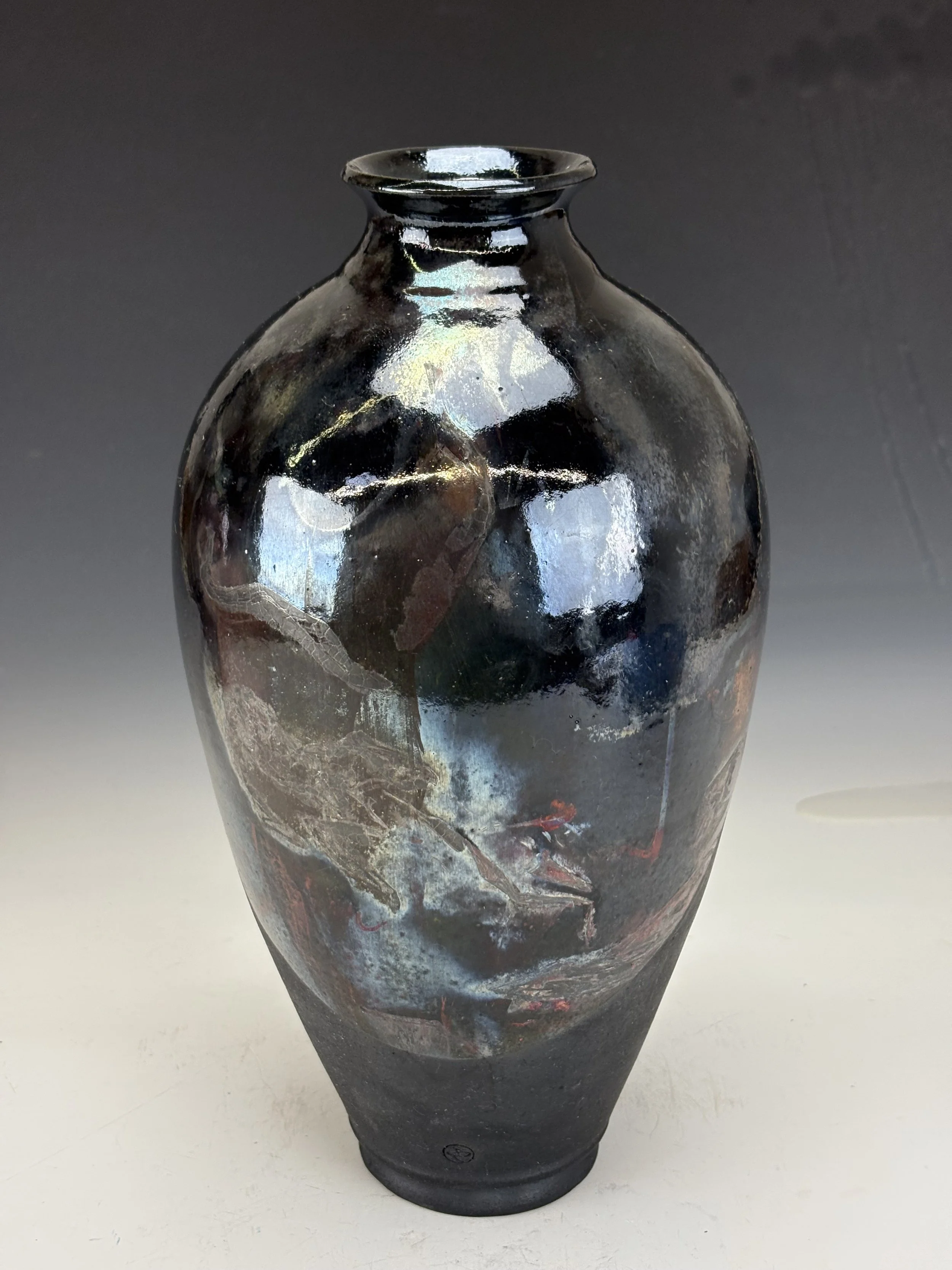 A tall, black ceramic vase with a glossy finish and abstract multicolored patterns in red, white, and earthy tones, set against a plain gray background.