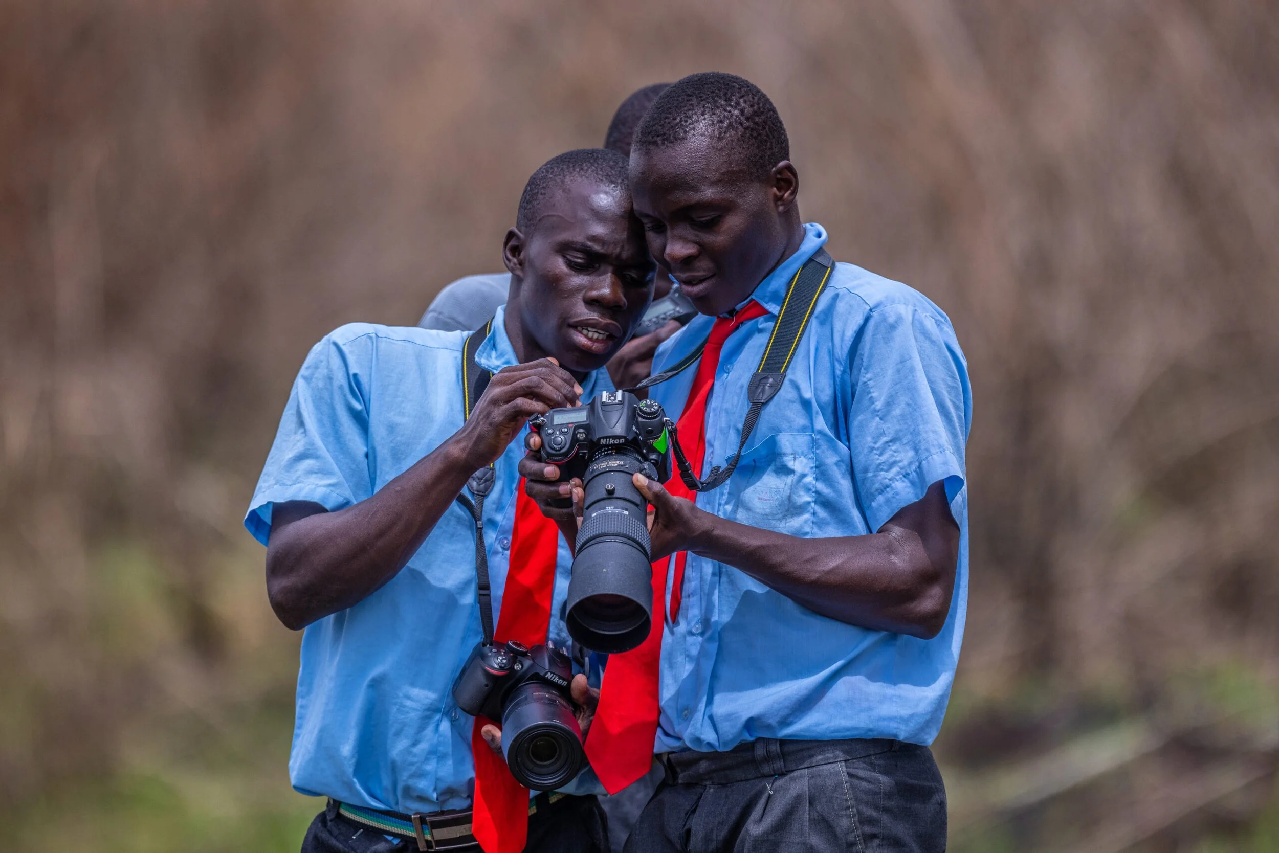 Two young men in blue shirts look at a Nikon camera, with one holding it and the other leaning in, both focused intently on the camera's screen.
