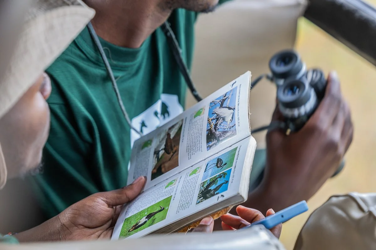 Two people, one holding a bird guidebook and the other holding binoculars, looking at the bird guidebook.