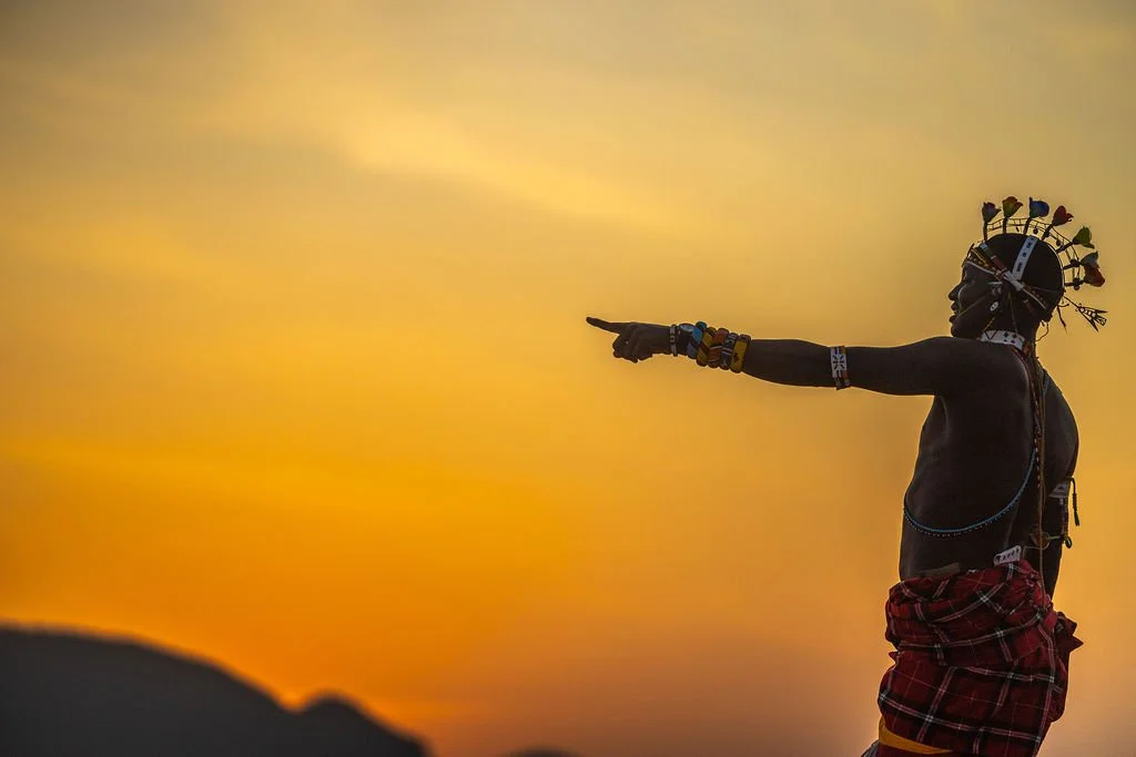 A person wearing traditional Maasai attire, including a headdress, jewelry, and a red shuka, standing against a colorful sunset sky, pointing into the distance.