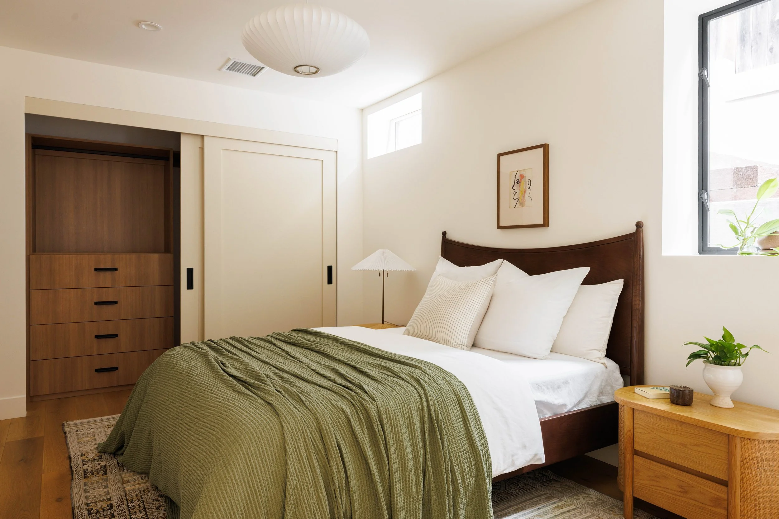 A cozy bedroom with a wooden bed frame, white bedding, green blanket, and pillow, a small side table with a plant and a glass cup, artwork on the wall, a window on the side letting in natural light, a built-in wardrobe with sliding doors, and a ceili