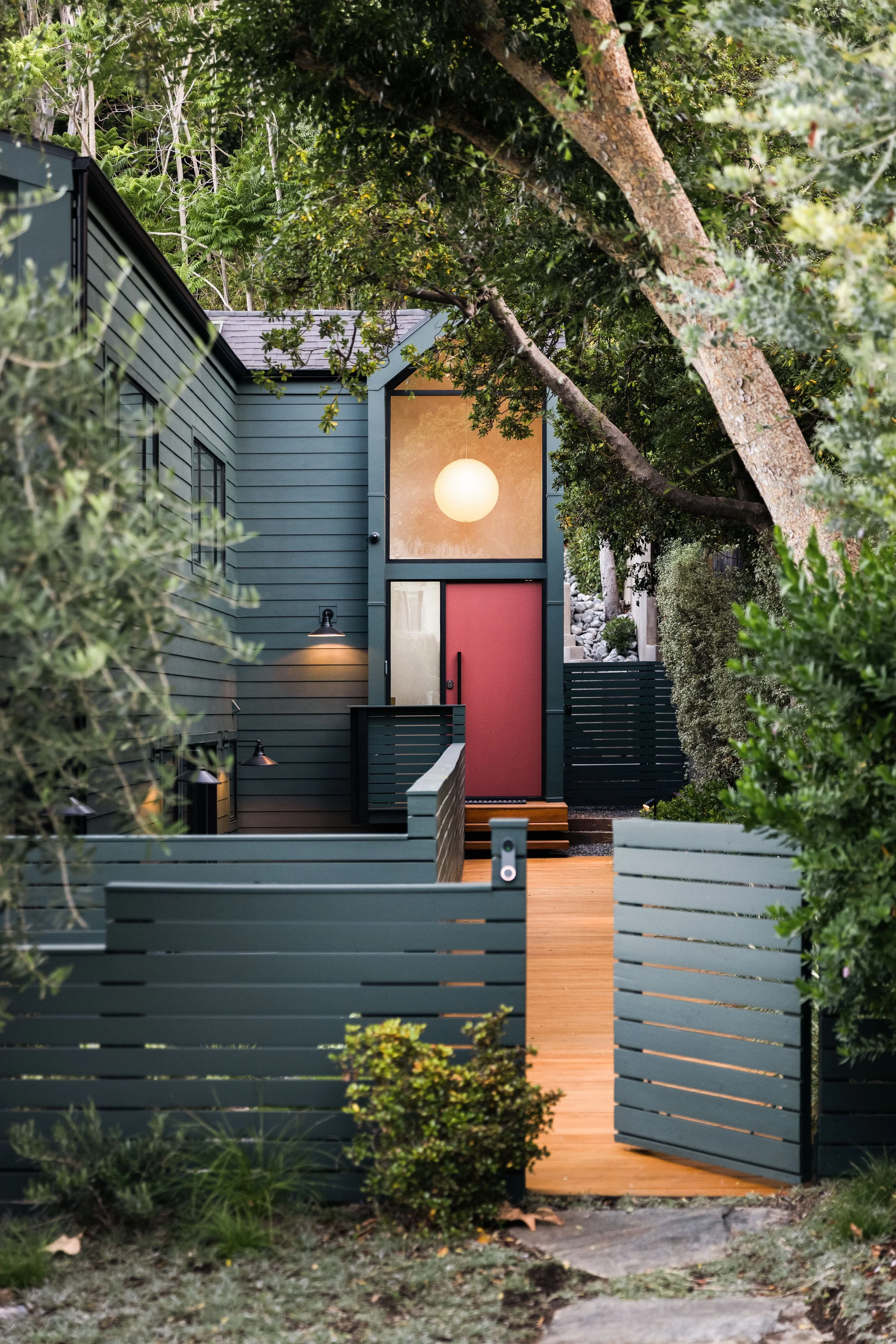 A cozy modern house with blue-gray siding and a red front door, surrounded by lush trees and greenery. A warm round light hangs in the front window, and a pathway with wooden steps leads to the entrance. A gray fence with horizontal slats borders the