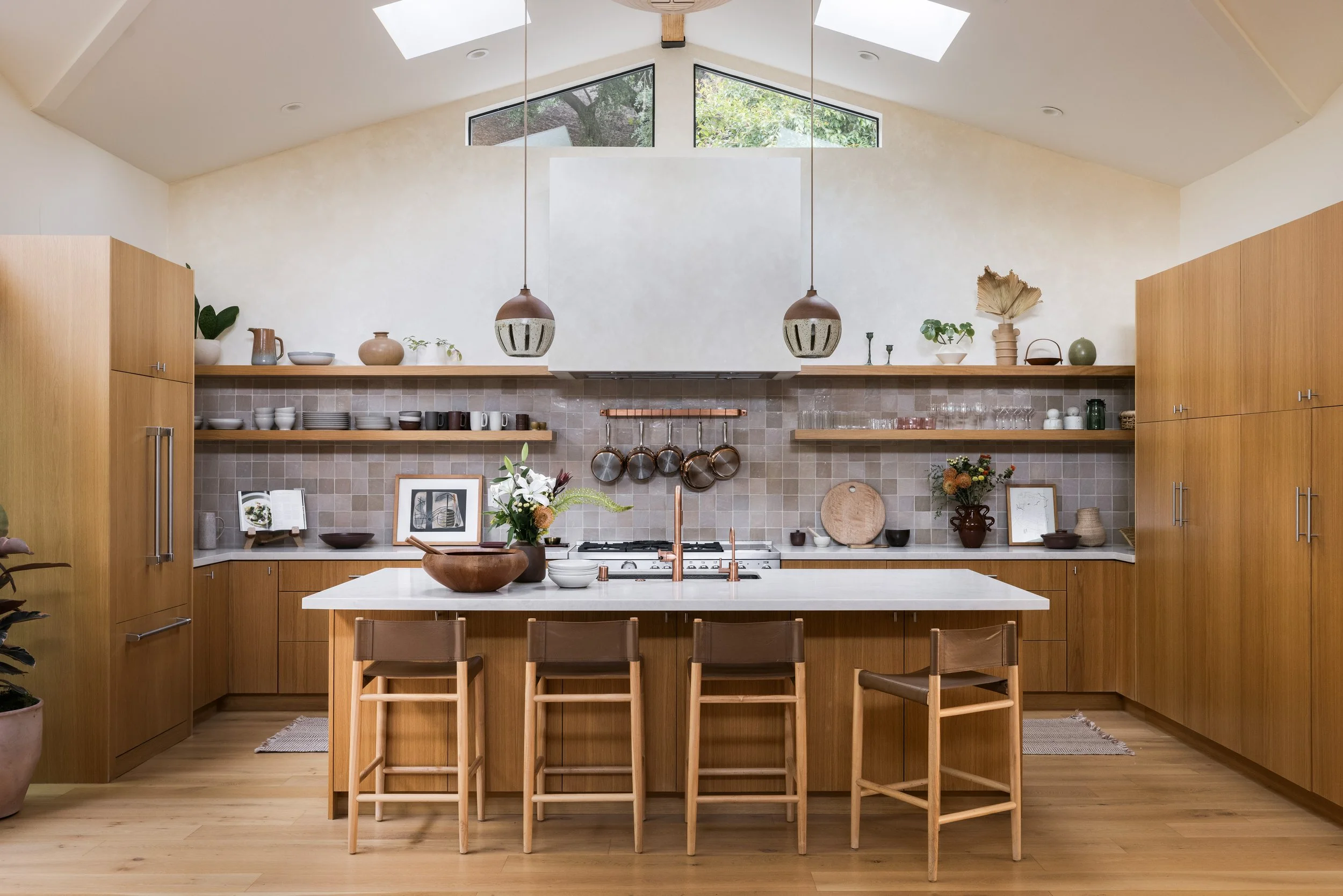 Modern kitchen with wooden cabinetry, open shelves with dishes and plants, a white island with a wooden bowl, and two pendant lights hanging from a high ceiling with skylights.