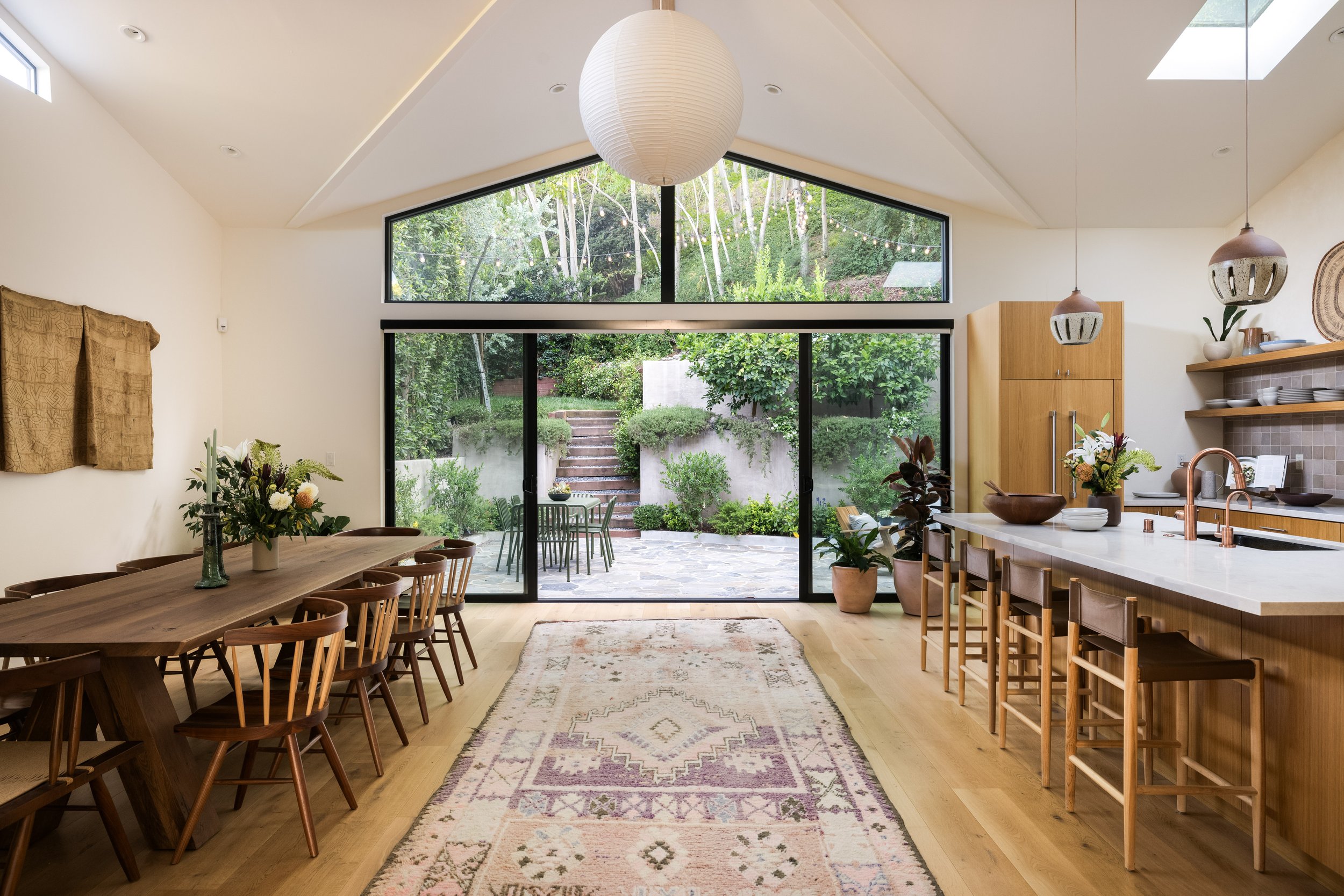 Open-concept kitchen and dining area with large windows showing outdoor greenery and string lights, wooden furniture, and a patterned area rug.