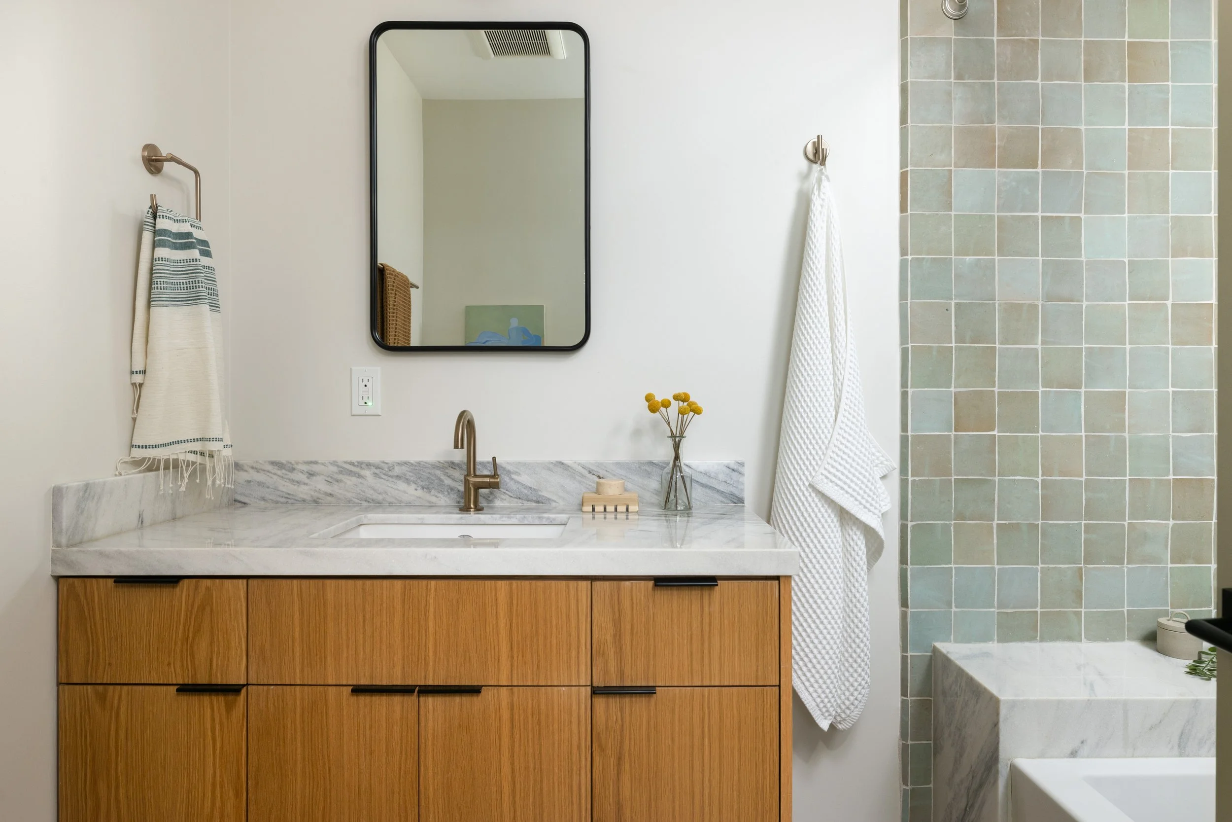 Bathroom vanity with wooden cabinet, marble countertop, and gold faucet. A mirror above the sink, with striped and white towels hanging on wall hooks. A small vase of yellow flowers on the counter. A tiled wall section to the right and a white towel 