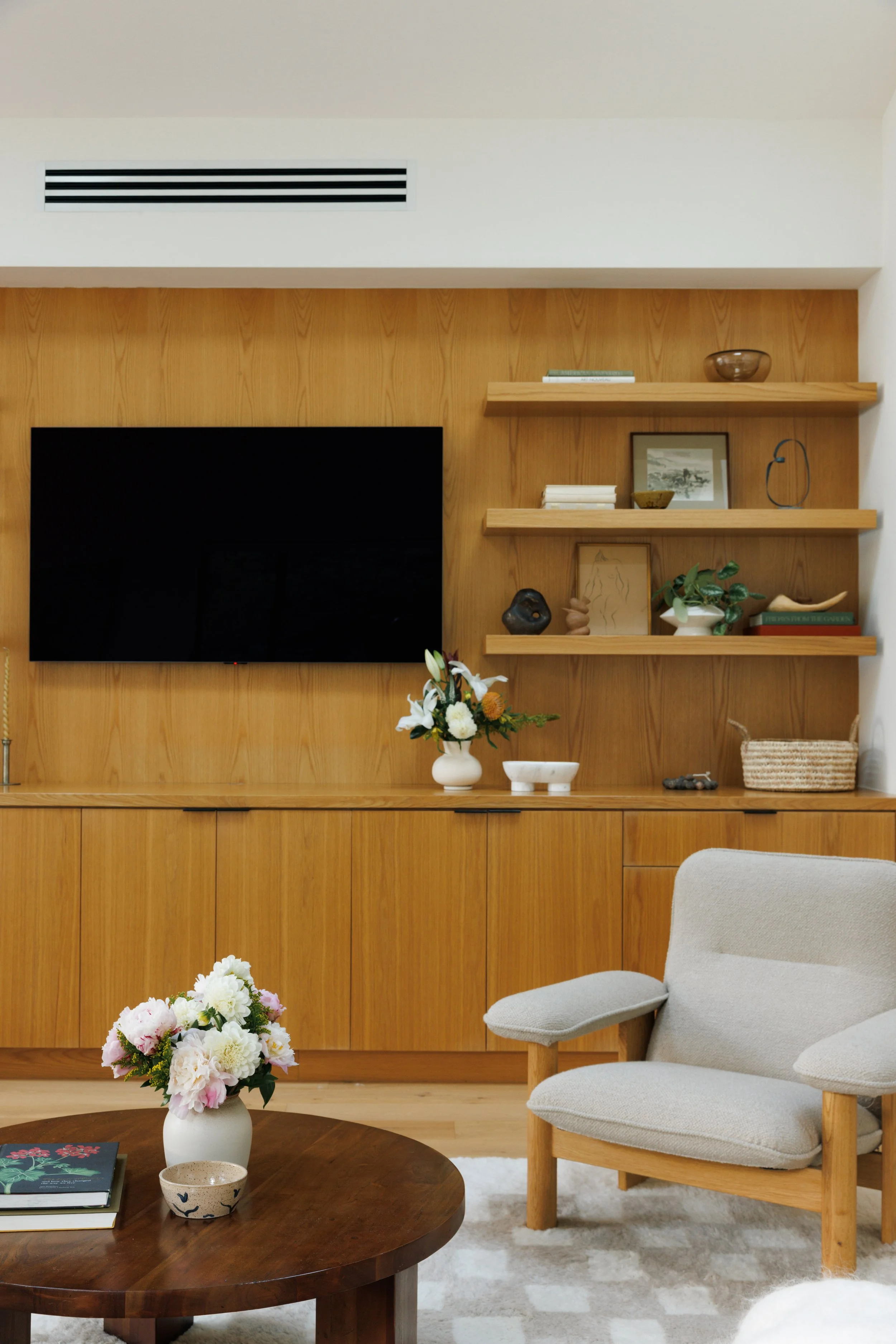 A cozy living room with a wooden wall featuring two shelves holding books, decorative items, and a framed picture. A flat-screen TV is mounted on the wall. There is an armchair with light-colored fabric and wooden legs, and a round wooden coffee tabl