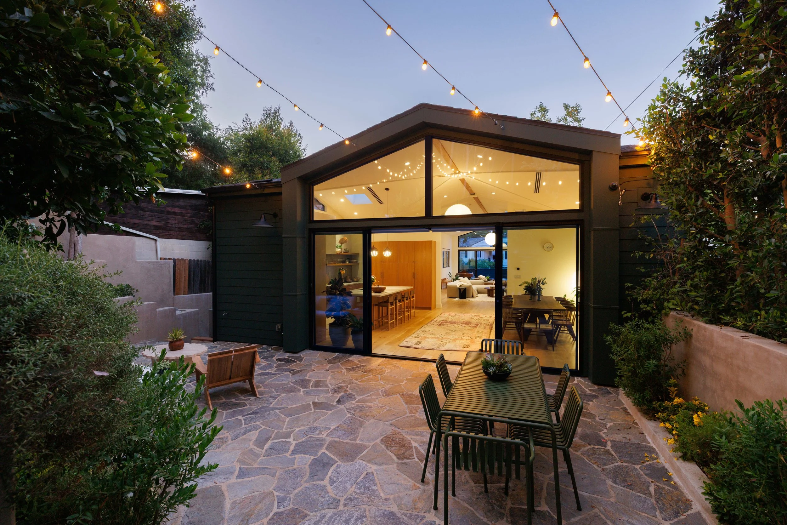 A backyard patio at dusk with string lights above. The patio has a stone floor, a dining table with chairs, and a view into a modern house with large glass windows showing a warmly lit interior living area.