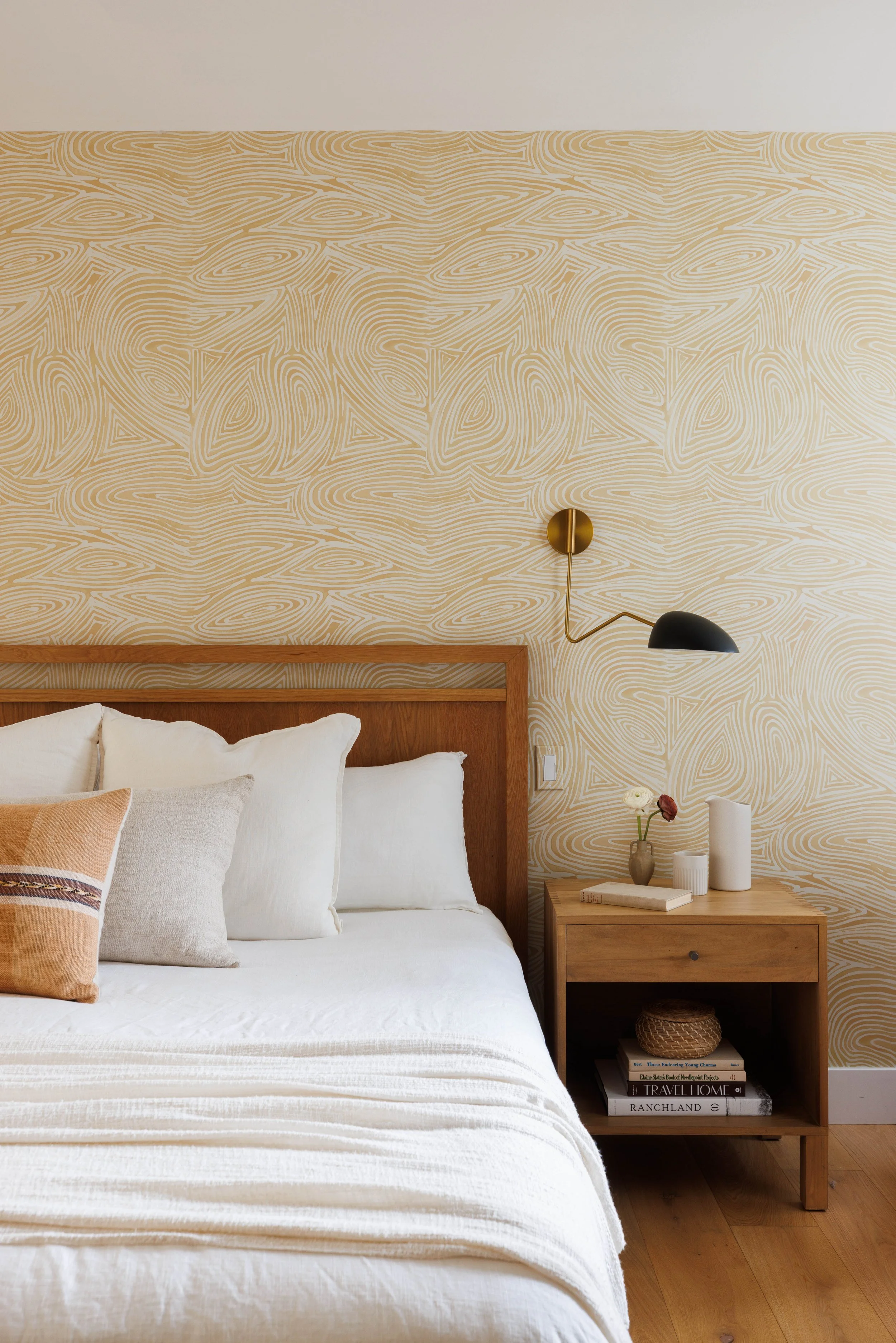 Bedroom with a white bed, beige patterned wallpaper, and a wooden nightstand holding white vases, a book, and a wicker basket, illuminated by a modern wall-mounted lamp.
