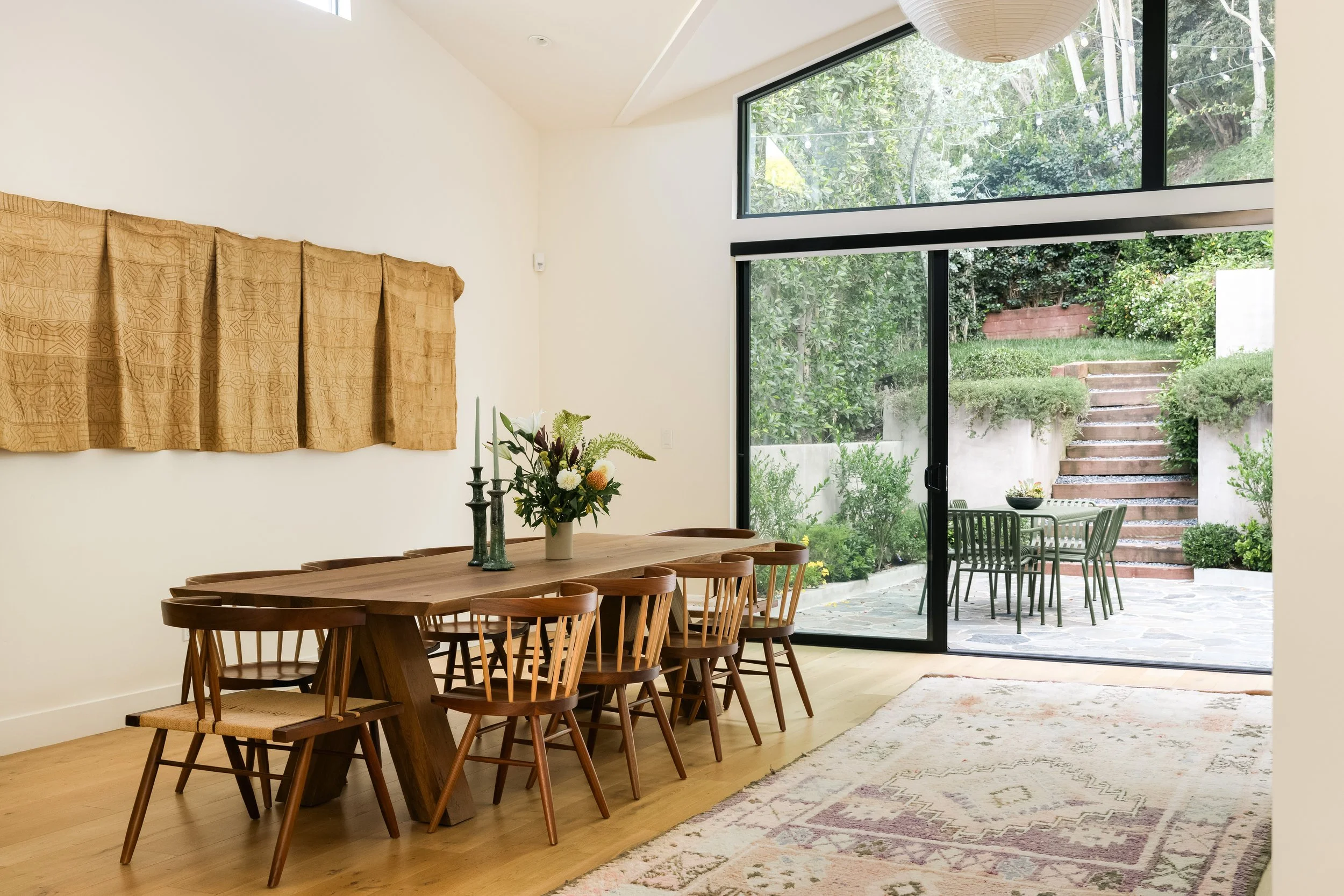 A dining area with a wooden table, chairs, a centerpiece with flowers, and large glass doors leading to an outdoor patio with greenery.