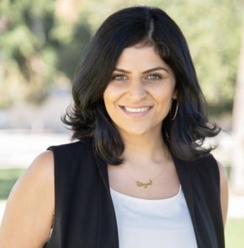 Listing agent Elga Kazanci, a woman with shoulder-length black hair, wearing a sleeveless black top with a white shirt underneath, gold jewelry, and smiling outdoors in a park with trees in the background.
