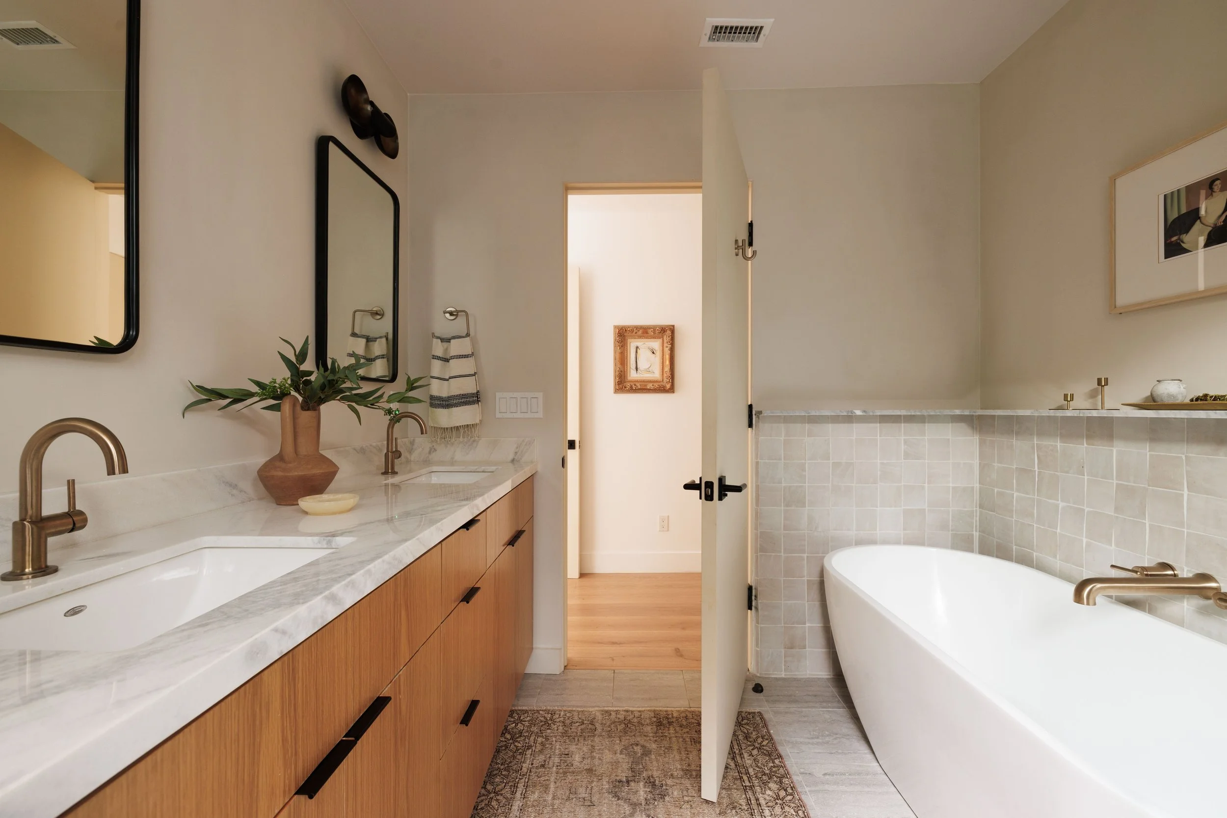A modern bathroom with a white bathtub, wooden vanity with a marble countertop, a sink, gold faucet, a mirror, framed pictures, a potted plant, and a towel hanging on a hook.
