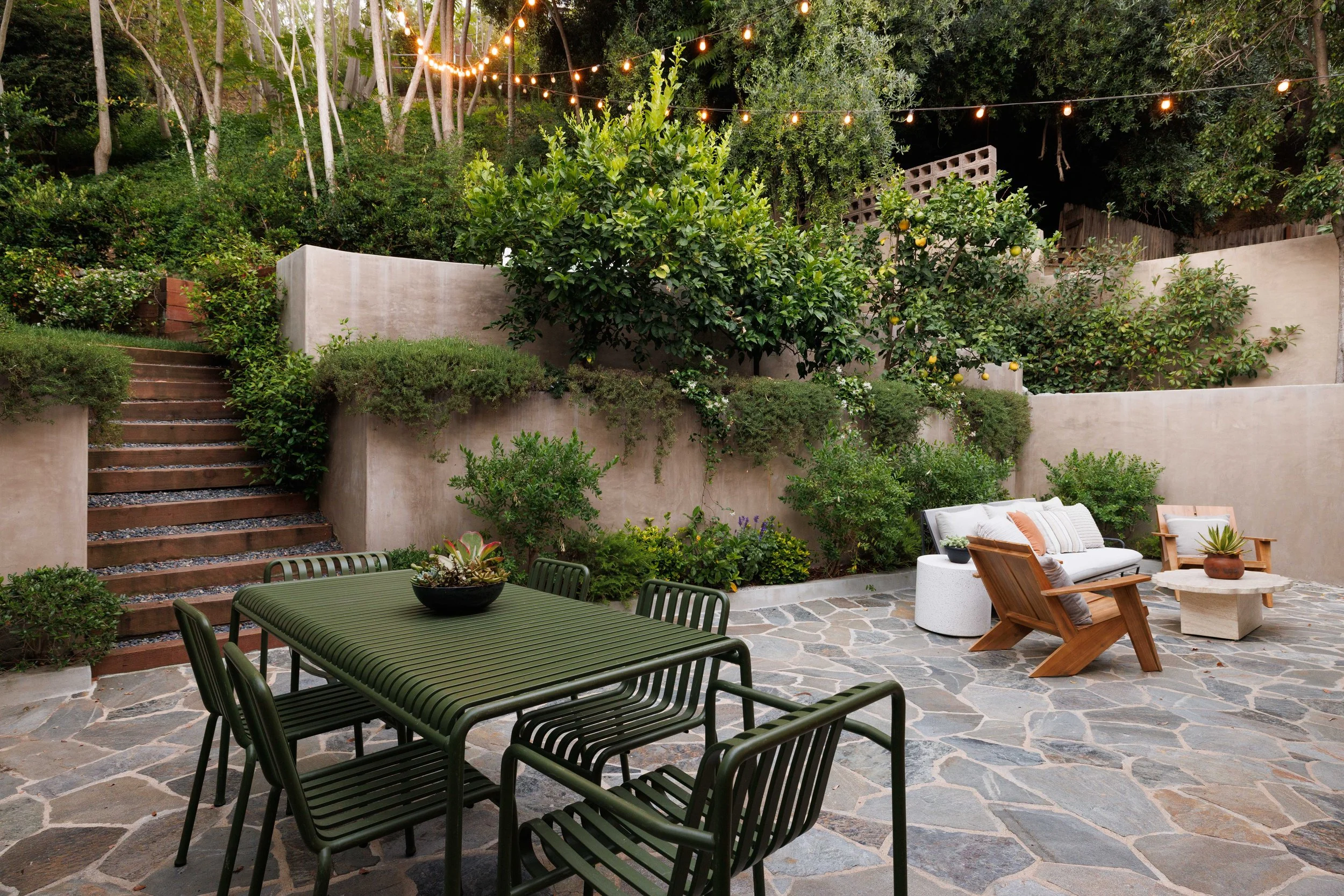 Outdoor patio area with string lights, greenery, trees, concrete walls, and outdoor furniture including a dark green table, chairs, a white cushioned lounge with pillows, and a wooden chair.