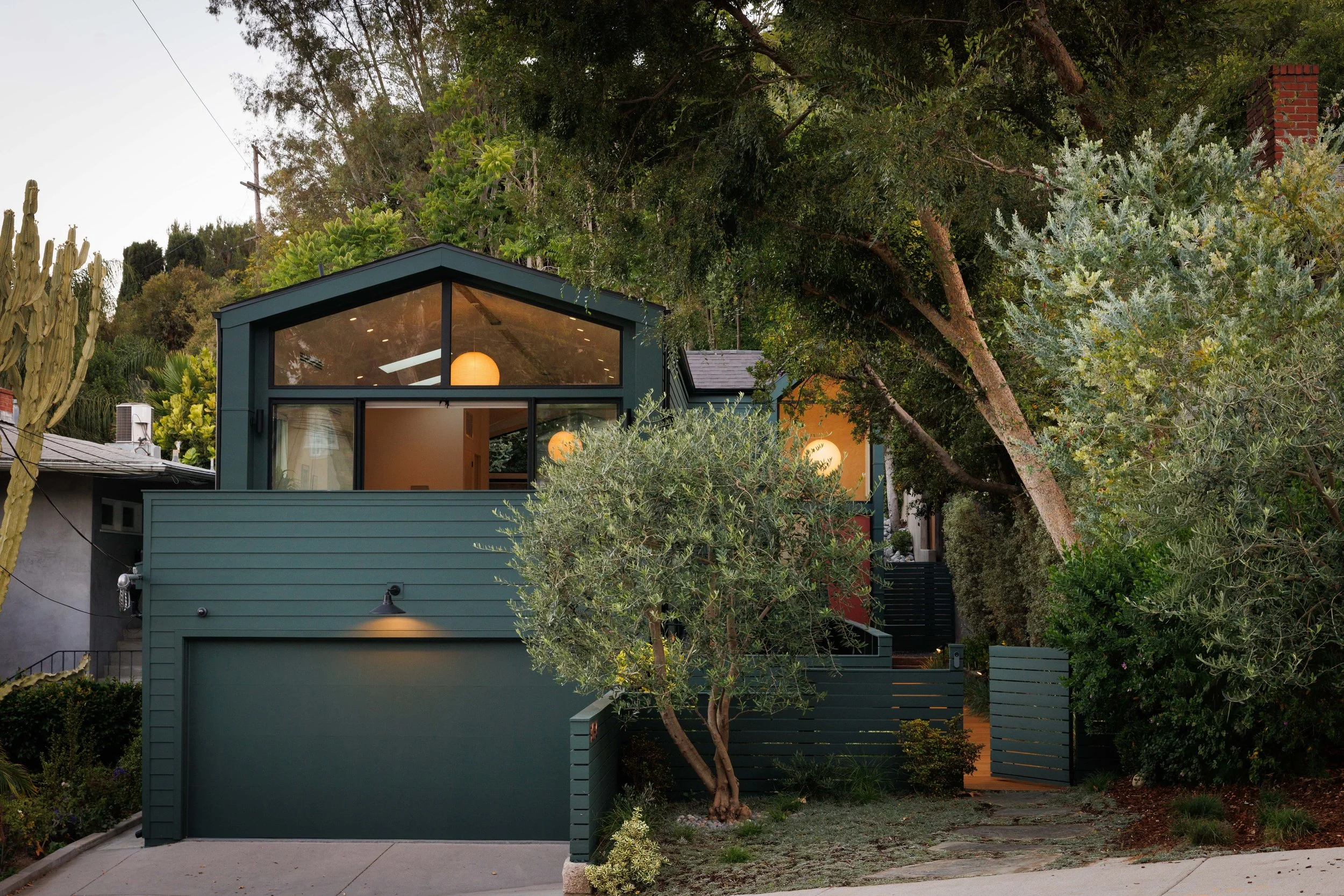 A modern two-story house at 3036 Castle Street in Silver Lake, with teal-colored siding and large glass windows, surrounded by trees and greenery in a suburban neighborhood at dusk.