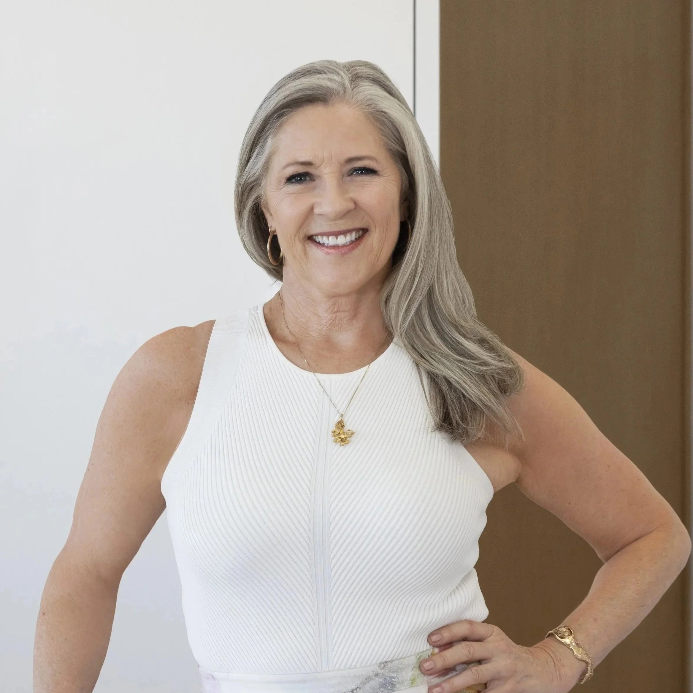 Listing agent Tracy Macrum, a smiling woman with long hair, wearing a white sleeveless top, gold hoop earrings, and a gold necklace with a pendant. She is standing indoors with a white wall in the background.