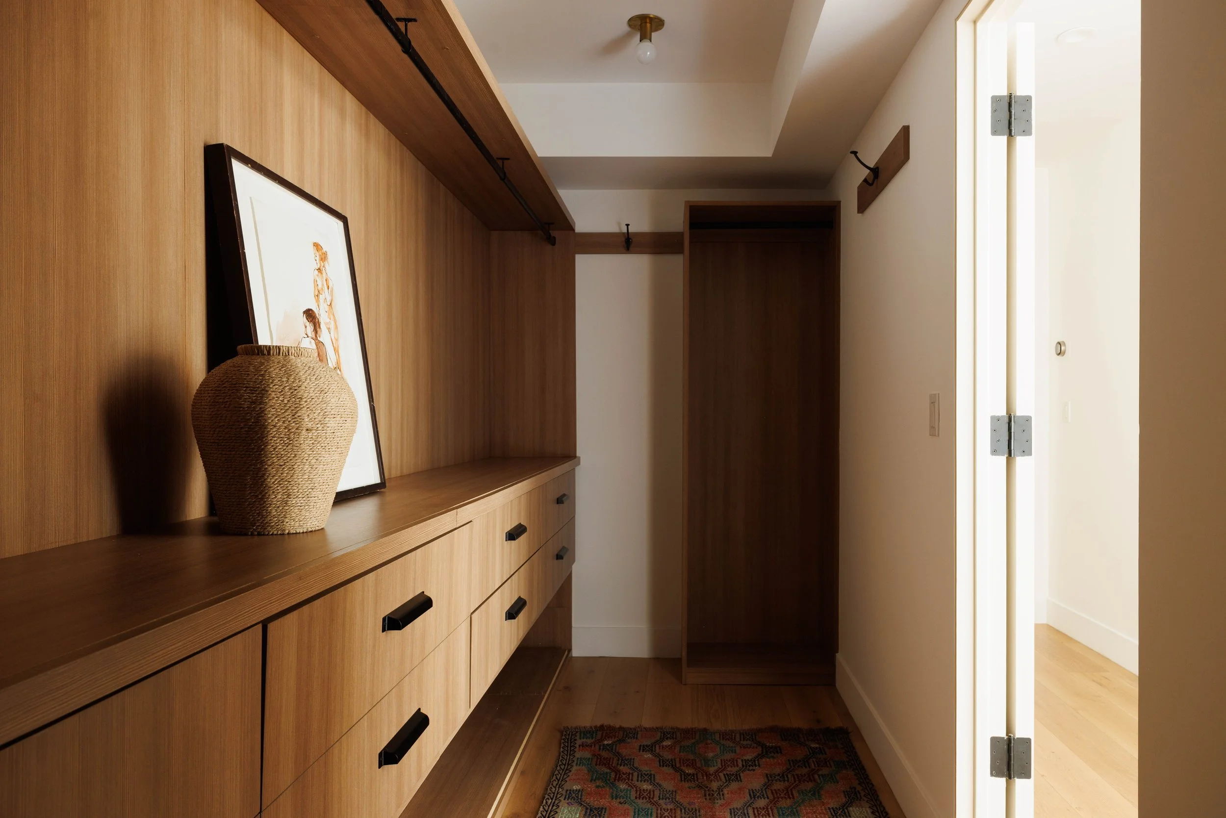 A narrow hallway with a wooden sideboard decorated with a framed artwork and a woven basket. The hallway has a wooden interior and an open door on the right side, with a light bulb on the ceiling.
