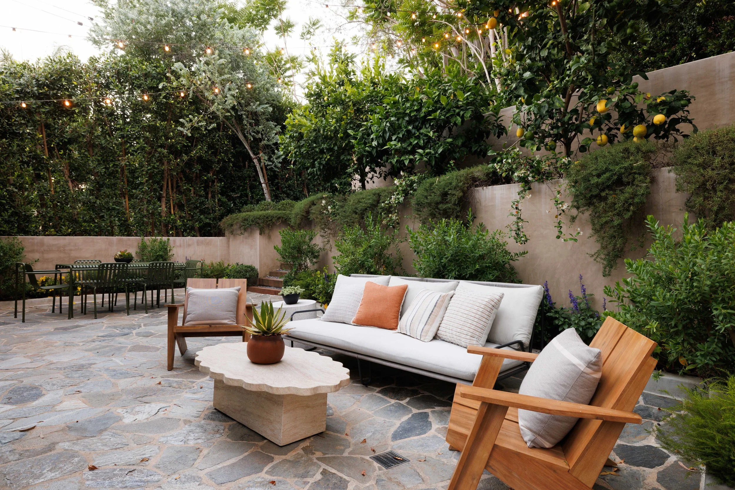 An outdoor patio with stone flooring, outdoor furniture including beige cushioned sofas with pillows, wooden chairs, a stone coffee table with a potted succulent, surrounded by green bushes, trees, and string lights.