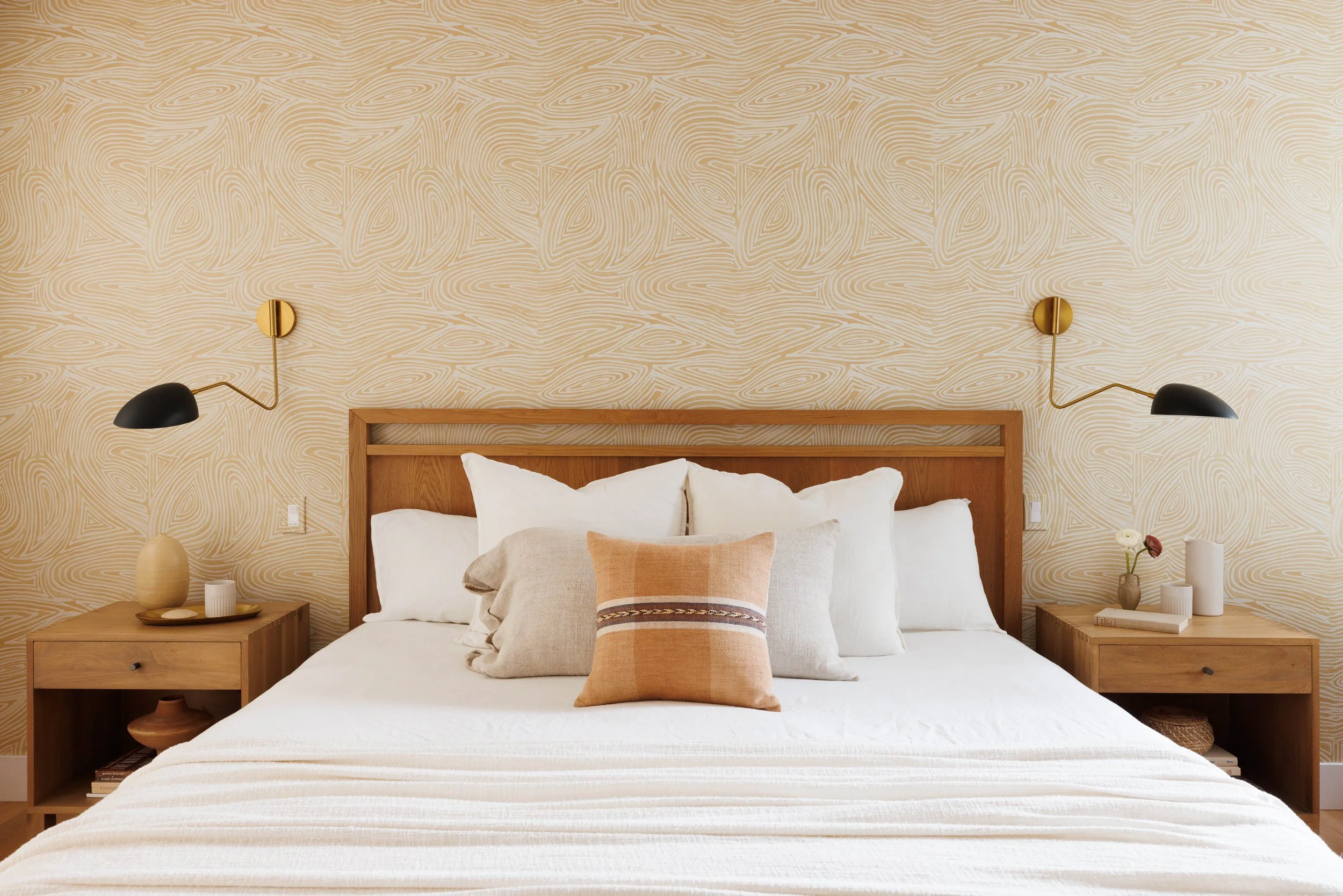 A neatly made bed with white linens, decorative pillows, and a wooden headboard. Two matching wooden nightstands with decorative vases and candles on each side, with wall-mounted black and gold reading lamps overhead. The wall has a beige abstract-pa