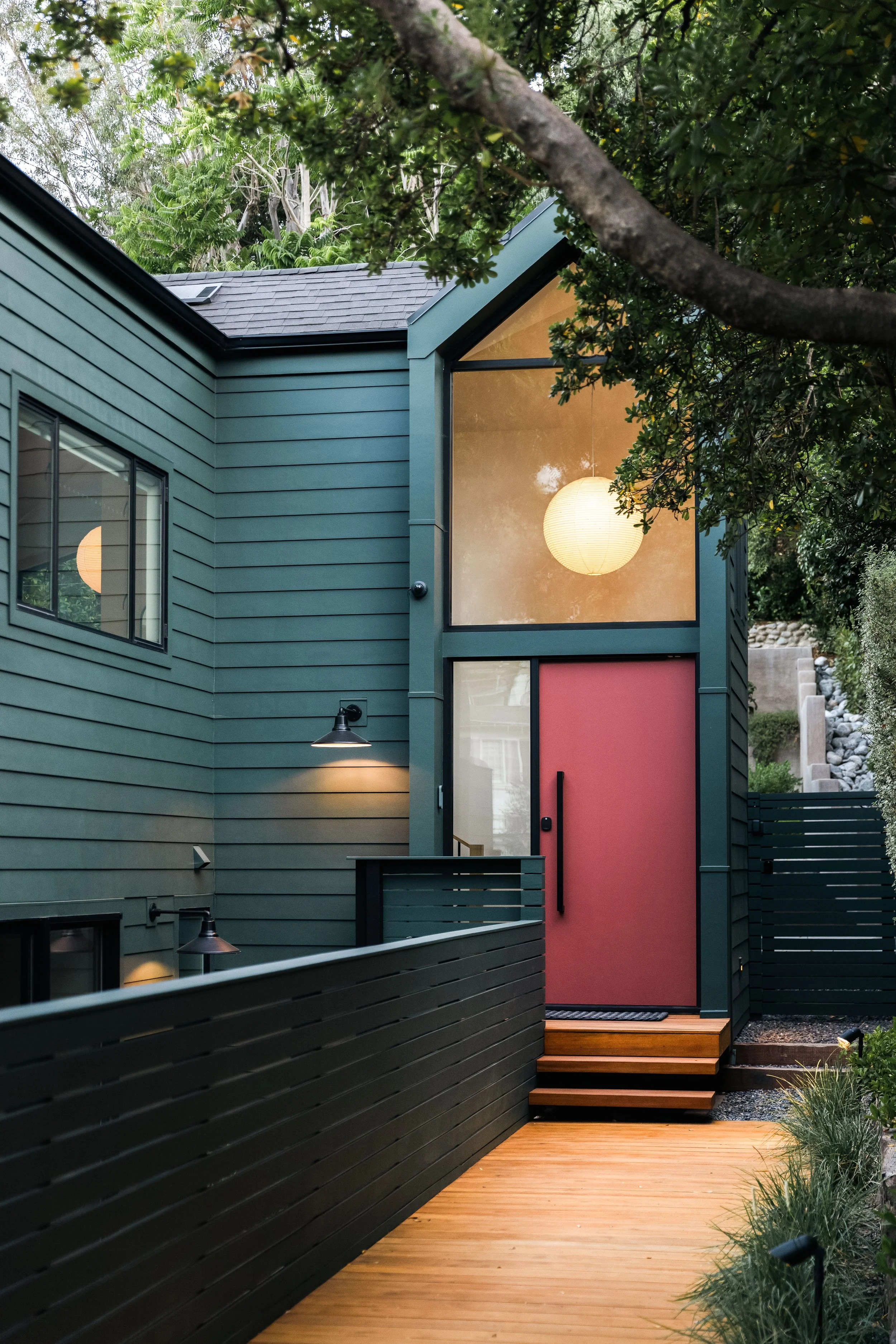 A modern house exterior with teal horizontal siding, large windows, and a red front door with black hardware. There is a small porch with wooden steps leading to the entrance, and a garden with plants along the pathway. A tree with green leaves parti