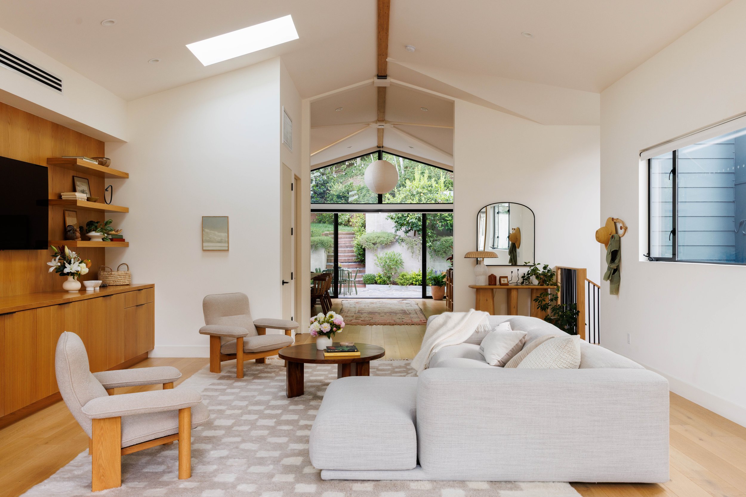 Living room with beige furniture, round wooden coffee table, light-colored rug, wooden wall shelves with decor, and large windows showing a garden.