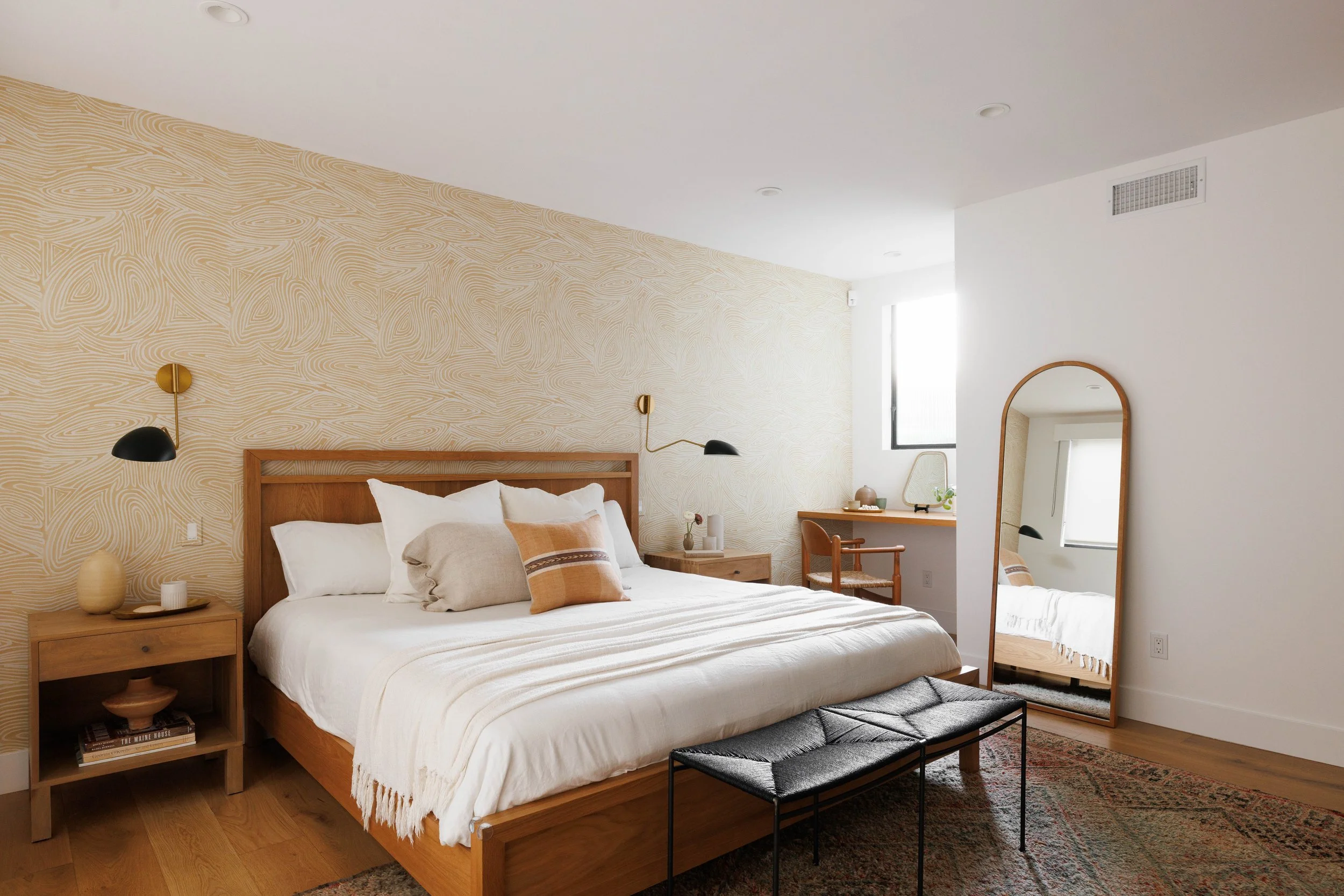 A modern bedroom with a wooden bed frame, white bedding, and decorative pillows. There are two black and gold wall-mounted lamps on either side of the bed. A wooden nightstand with a vase and books is beside the bed. A large mirror with a wooden fram