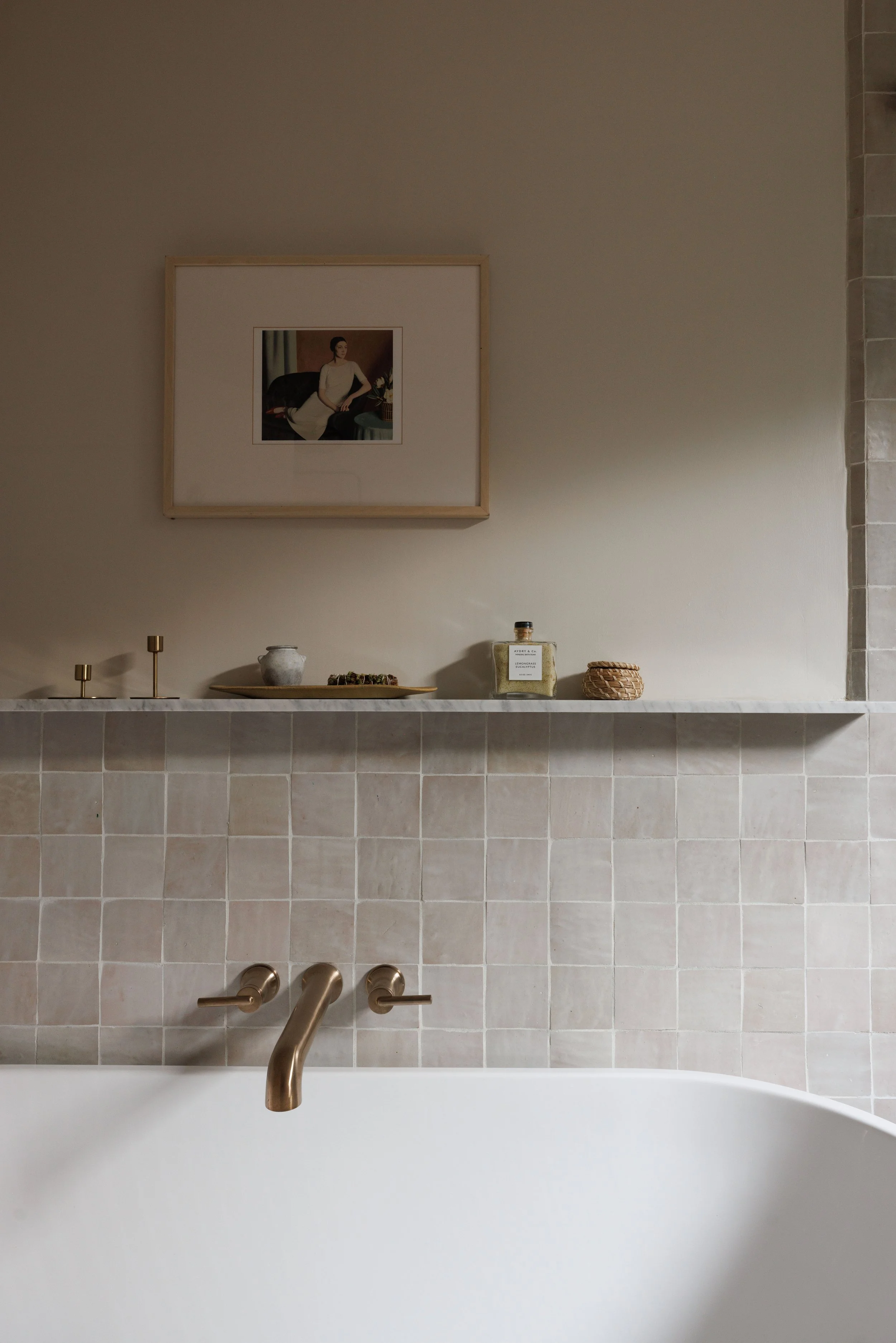 Part of bathroom with beige tiled wall, gold-colored bathtub fixtures, decorative objects on a marble shelf, framed picture of a woman in a white dress, and a bathtub in the foreground.