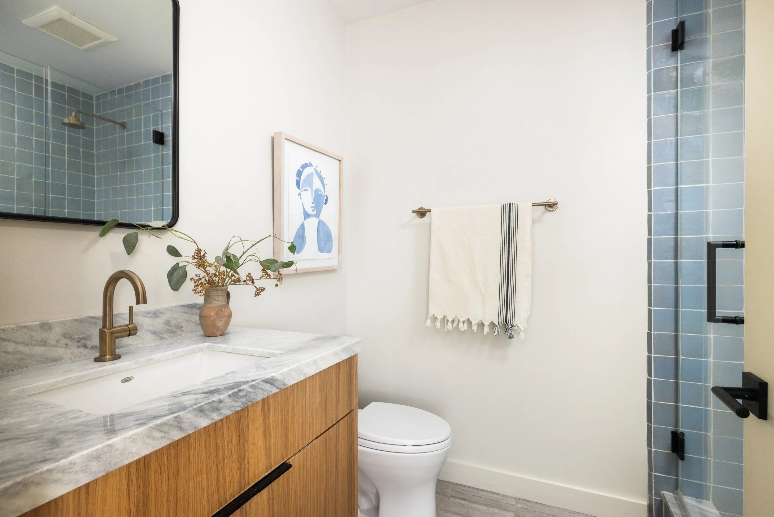 A modern bathroom featuring a marble countertop with a brass faucet, a framed abstract art piece, a plant in a small vase, a white towel hanging on a silver bar, a toilet, and a shower area with blue tiles and a black shower head.
