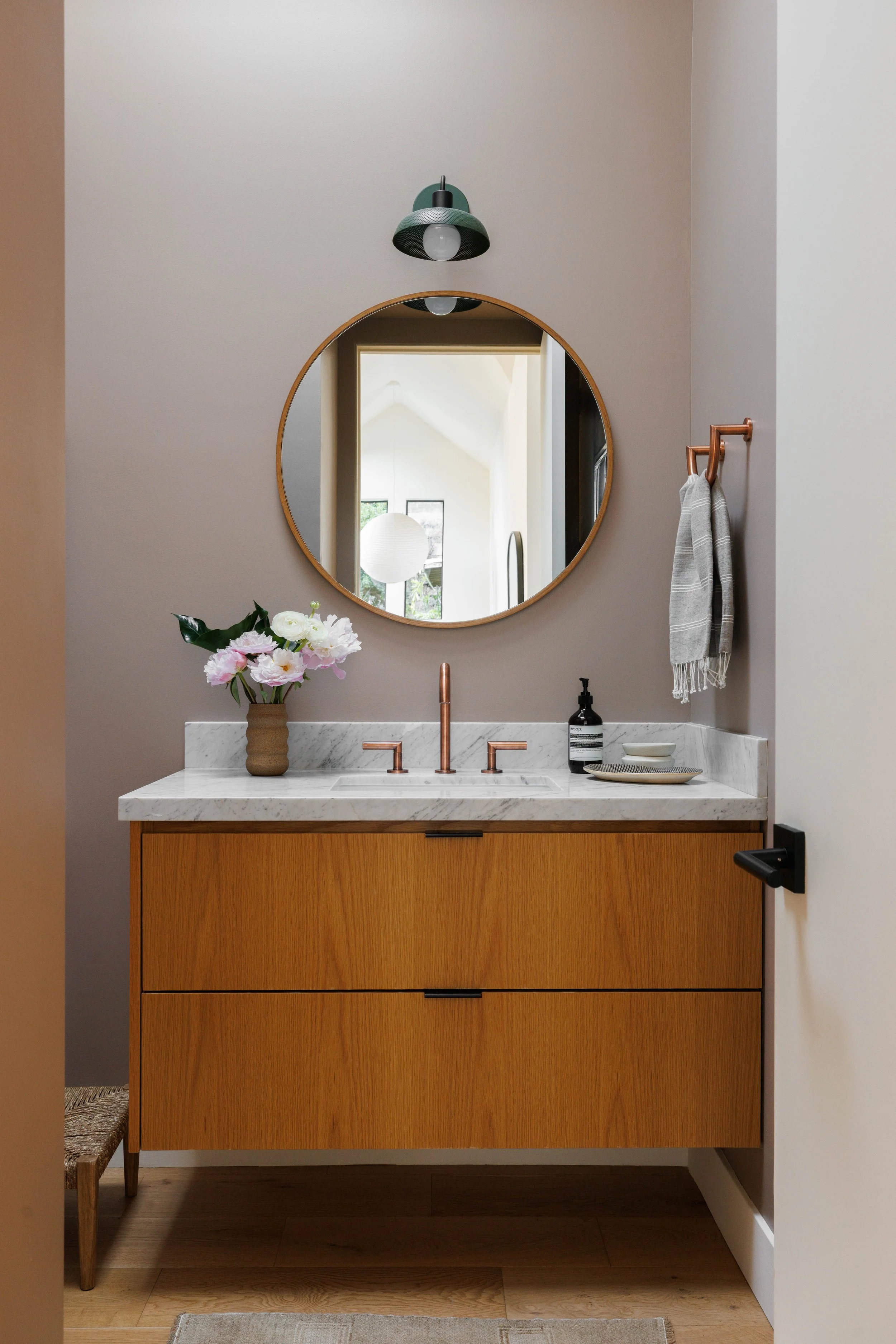 A bathroom vanity with a white marble countertop, a round mirror with a wooden frame, a black soap dispenser, a bowl, a vase with pink and white flowers, and a tan towel hanging on a copper towel ring. A green wall sconce with a bulb is mounted above