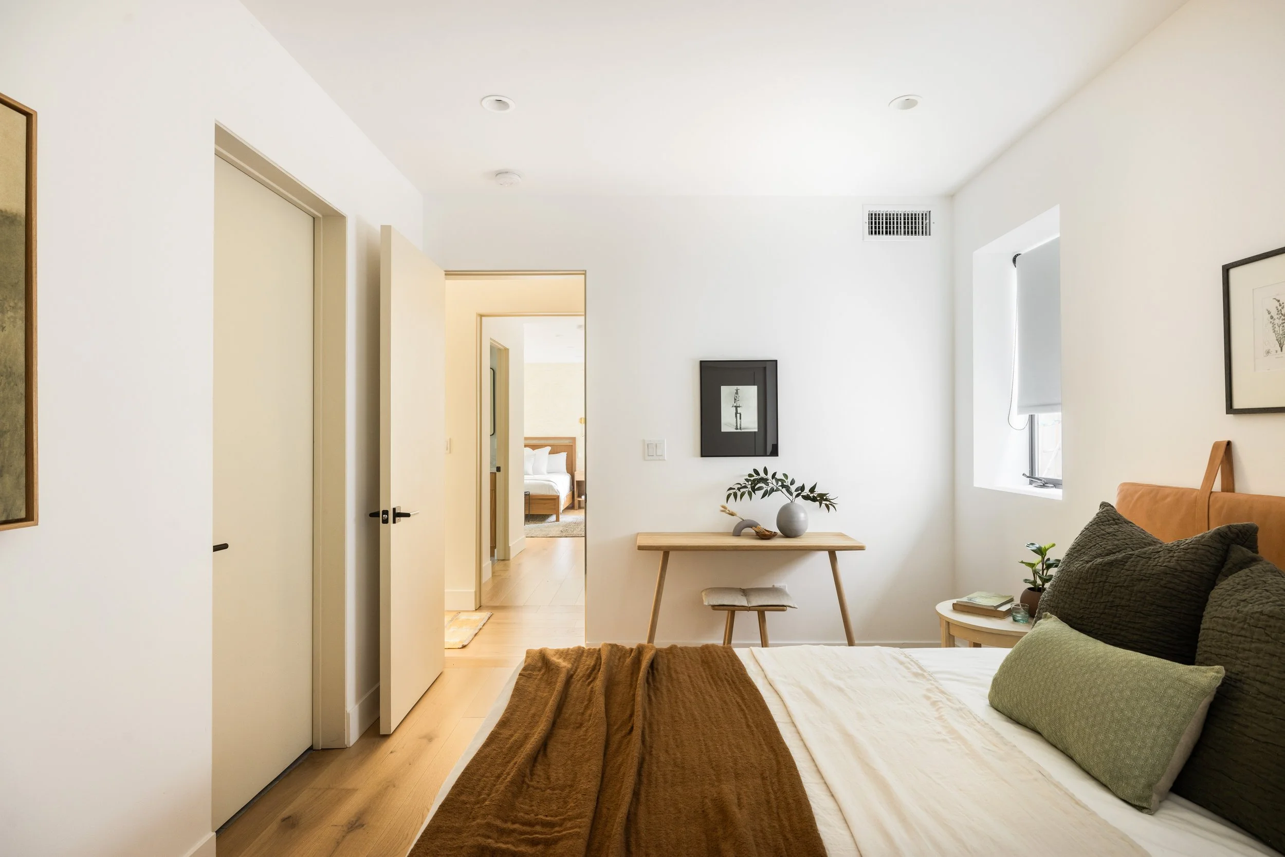 Bright, minimalist living room with a bed in the foreground, a small wooden table with a vase and plant, framed artwork on the walls, a window with white blinds, and a door leading to a bedroom with a bed visible.
