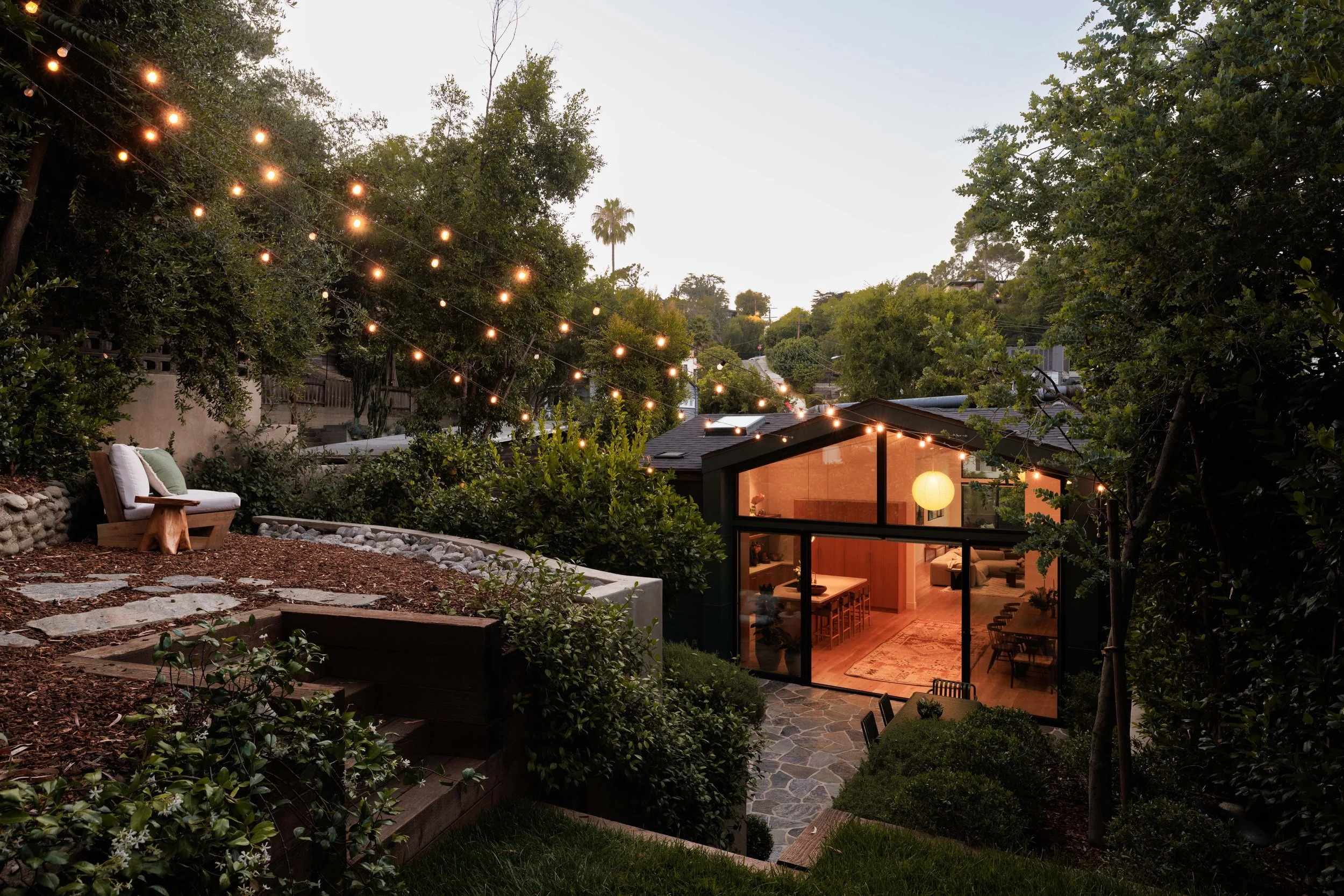 A cozy backyard with string lights hanging overhead, a white outdoor sofa with cushions on a stone and mulch patio, surrounded by lush trees and plants, and a modern glass-walled house with warm interior lighting.
