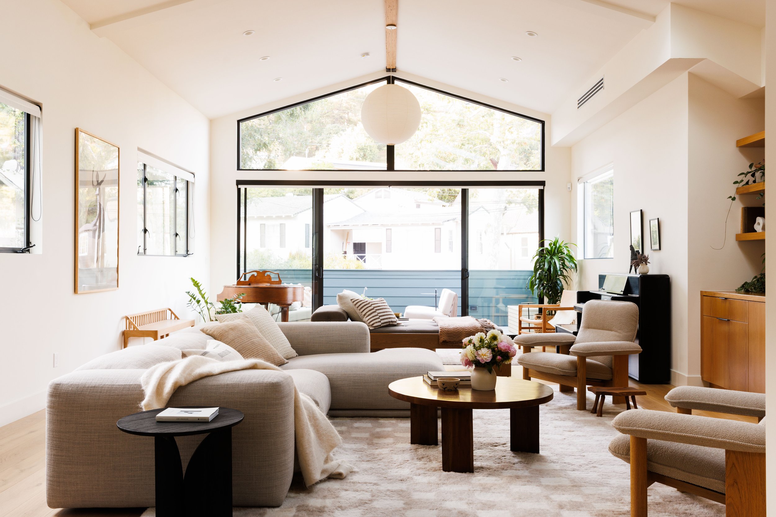 Living room with large windows, beige sofa with pillows, round wooden coffee table with flower arrangement, additional seating, and wall art. Natural light and indoor plants are visible.