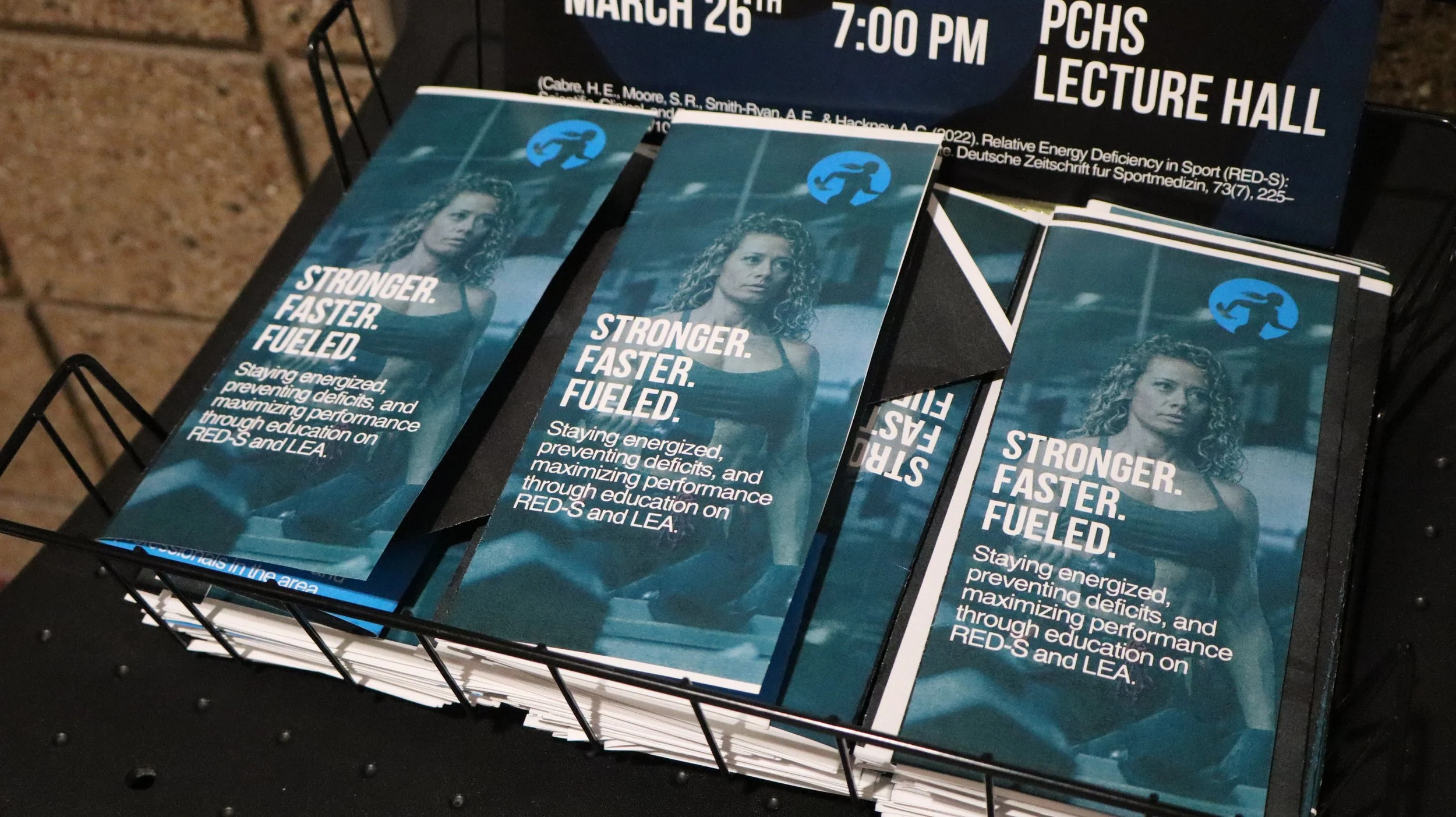 Brochures titled 'Stronger. Faster. Fueled.' featuring a woman in athletic attire, promoting education on energization, deficit prevention, and performance maximization, displayed in a black wire basket at an event.