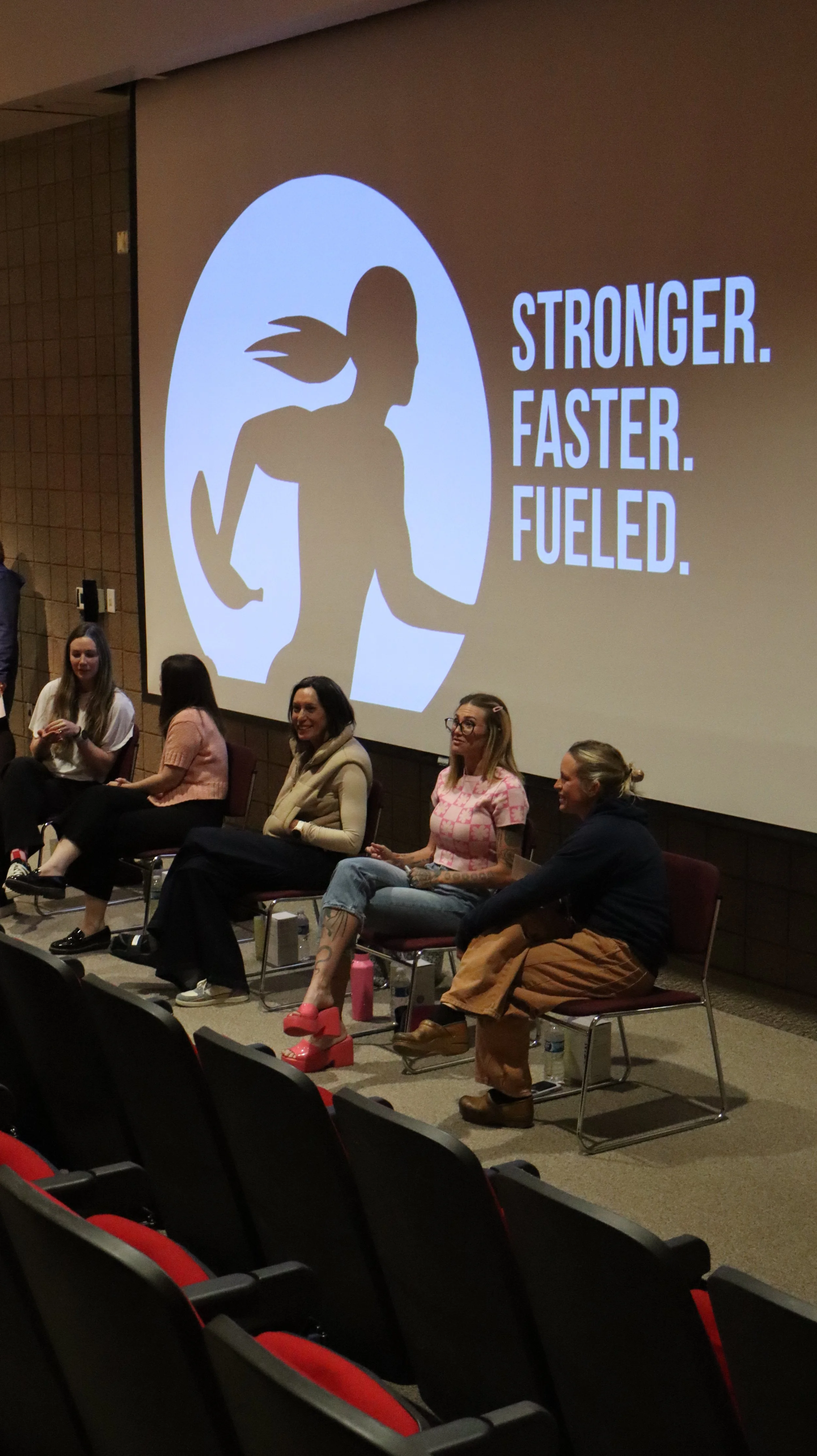 A group of women sitting on chairs in front of a large screen displaying a silhouette of a running woman and the words 'Stronger. Faster. Fueled.'