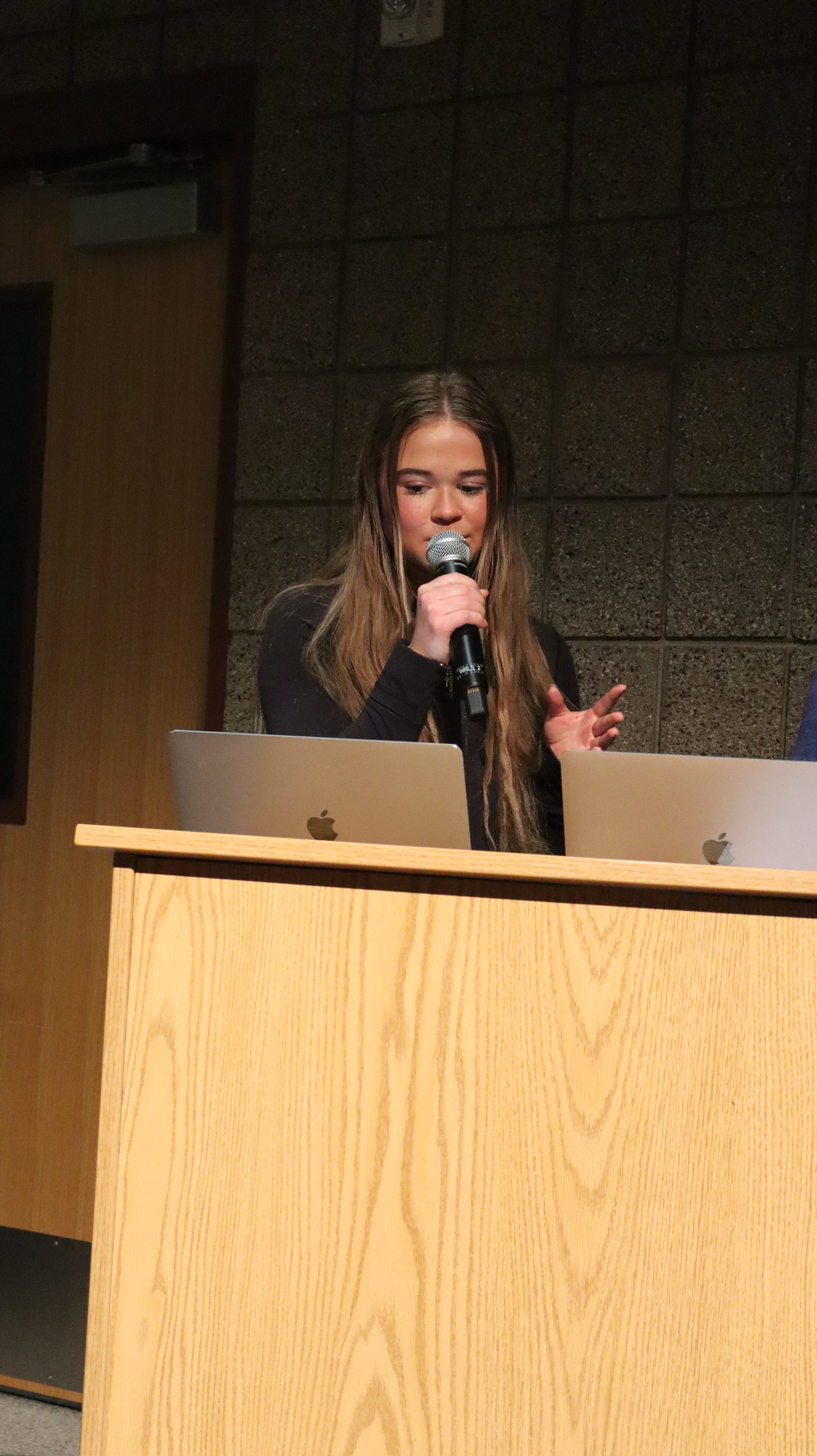 A young woman speaking into a microphone at a podium with two laptops in front of her.