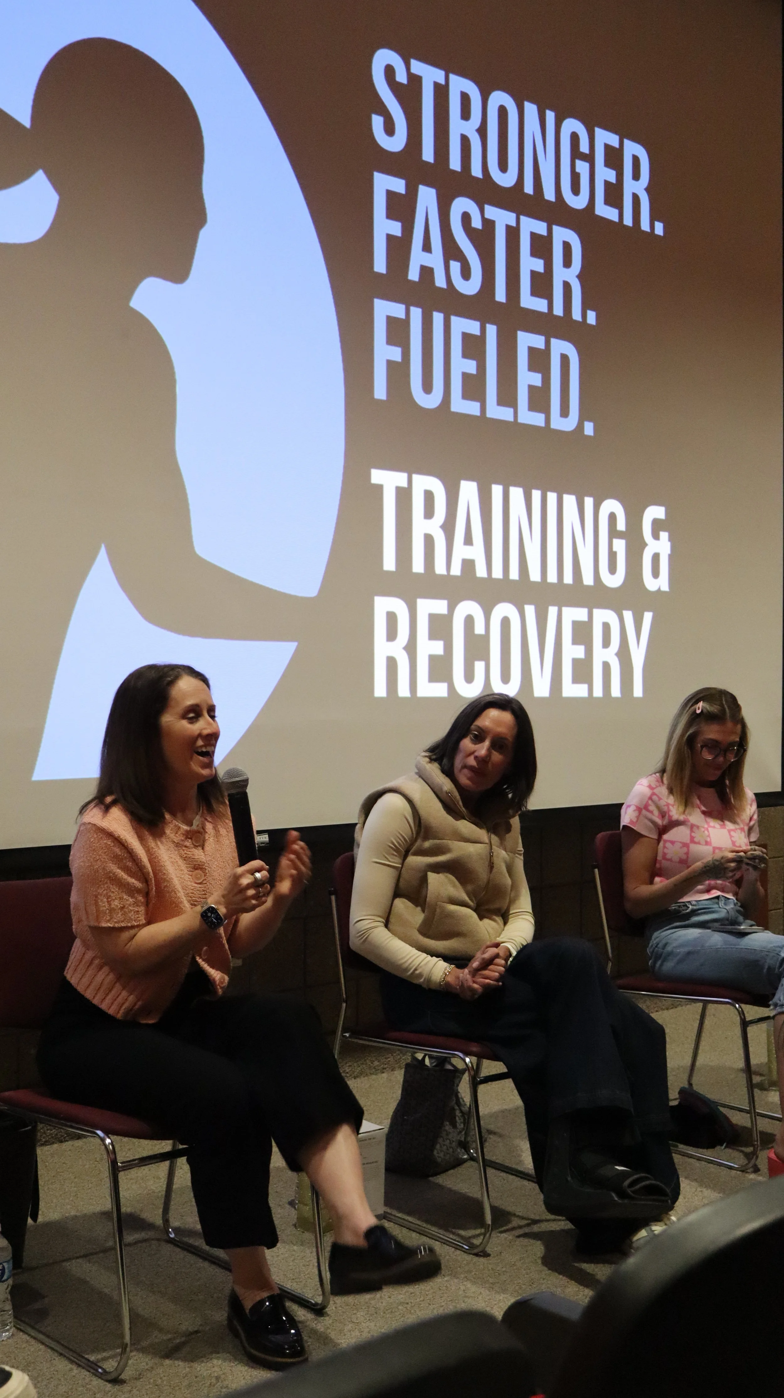 A woman is speaking into a microphone during a panel discussion in a conference room. Three women sit on chairs on stage, with a large screen behind them displaying text that reads: "Stronger. Faster. Fueled. Training & Recovery." The women are engaged and attentive, with one woman smiling and gesturing as she speaks.