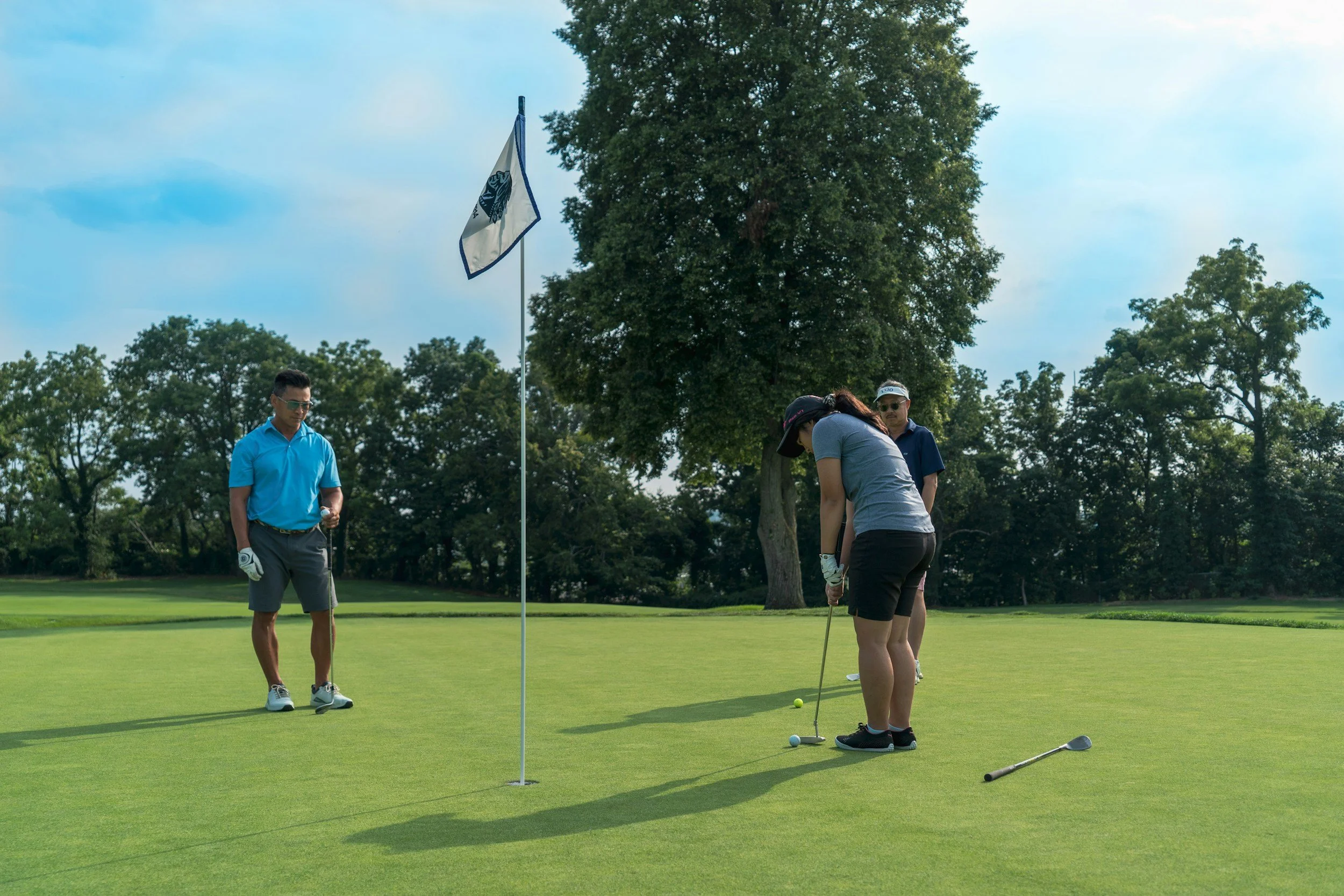 People playing golf on a green, with a flagstick and golf balls on the grass. Two women are preparing to putt, and a man is standing nearby watching. Large trees are in the background under a partly cloudy sky.