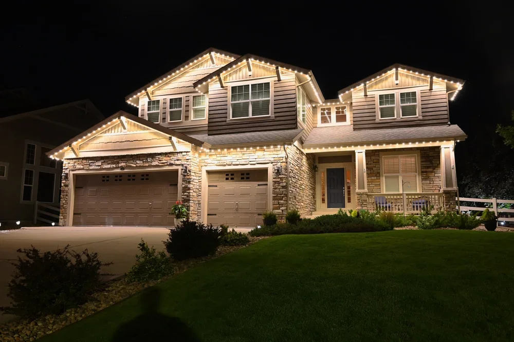 A two-story house illuminated at night with string lights outlining the roof and windows. It has a stone and siding exterior, a double garage, and a small porch with a railing. The front yard has well-maintained grass and shrubs.