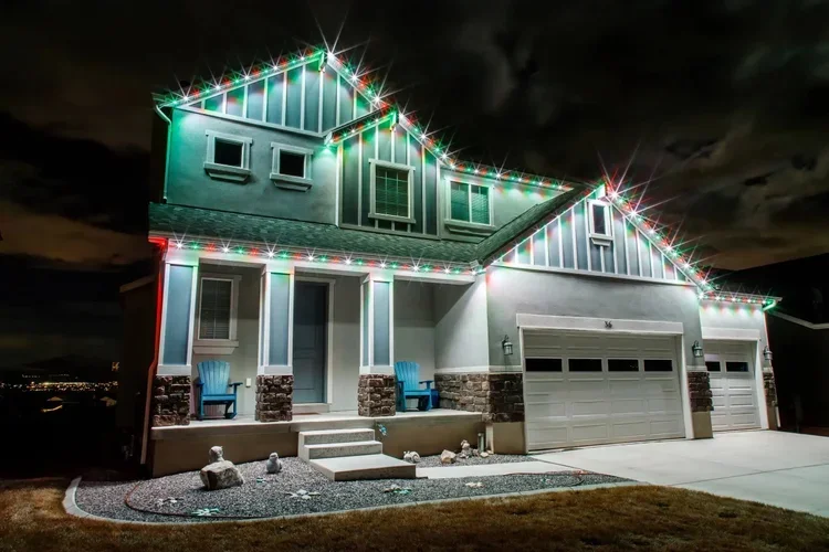A two-story house decorated with colorful Christmas lights at night, with two blue chairs on the front porch and a stone border surrounding a small yard.