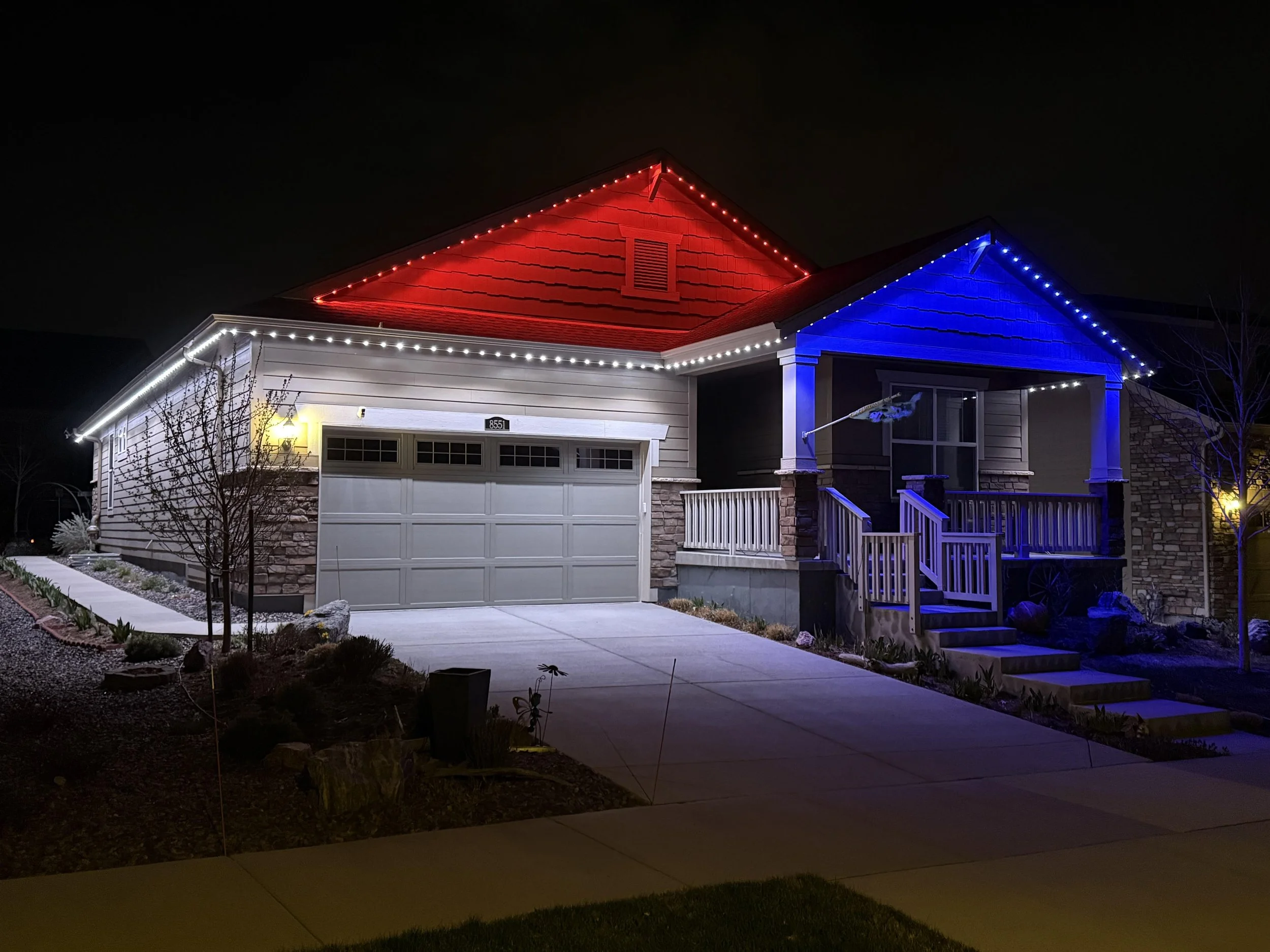 A house decorated with red, white, and blue Christmas lights at night, with a front yard and sidewalk visible.