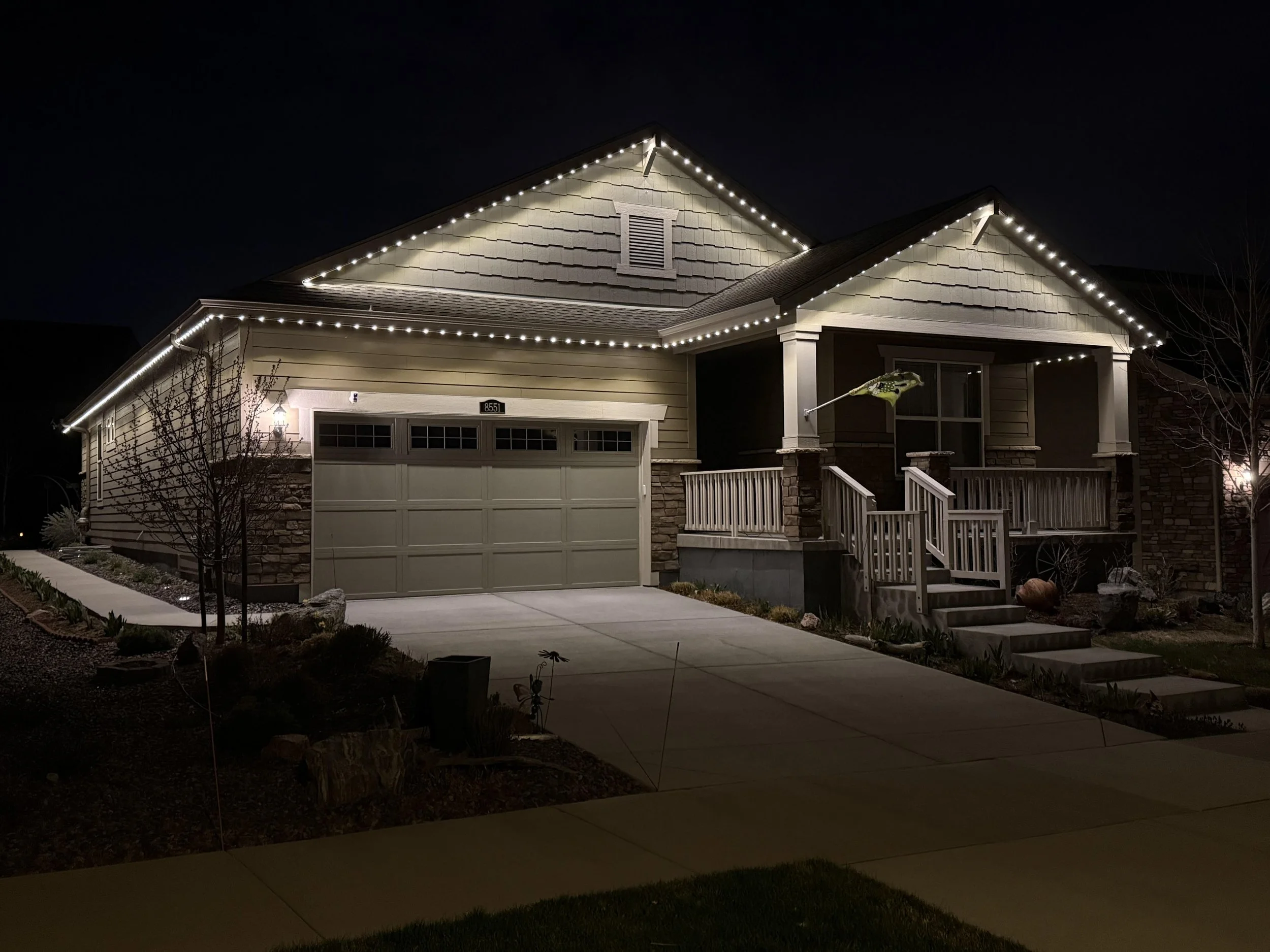 Nighttime photo of a house with outdoor string lights outlining the roof edges and front porch, a tree, and a sidewalk in the front yard.
