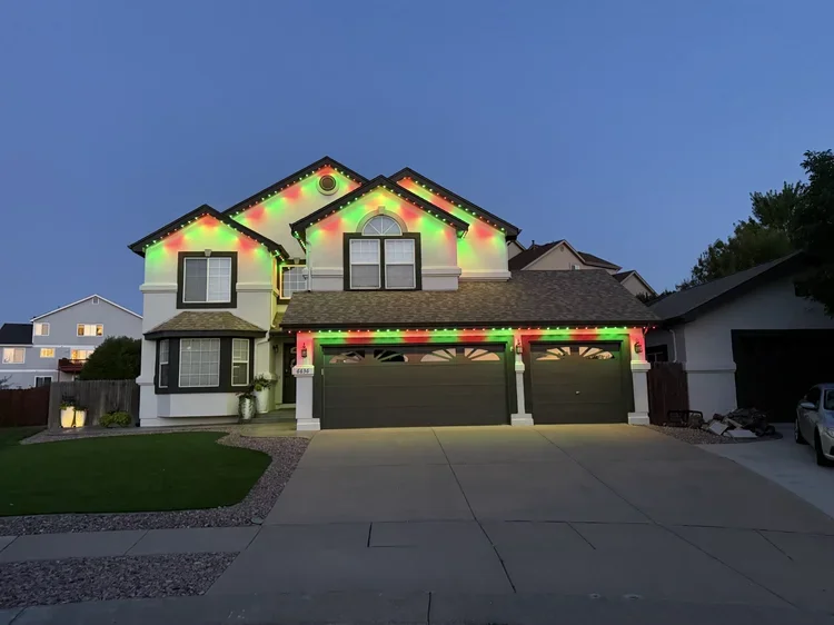 A large two-story house decorated with colorful Christmas lights along the roofline and around the garage at dusk. The house has a well-maintained lawn, a sidewalk, and neighboring houses nearby.