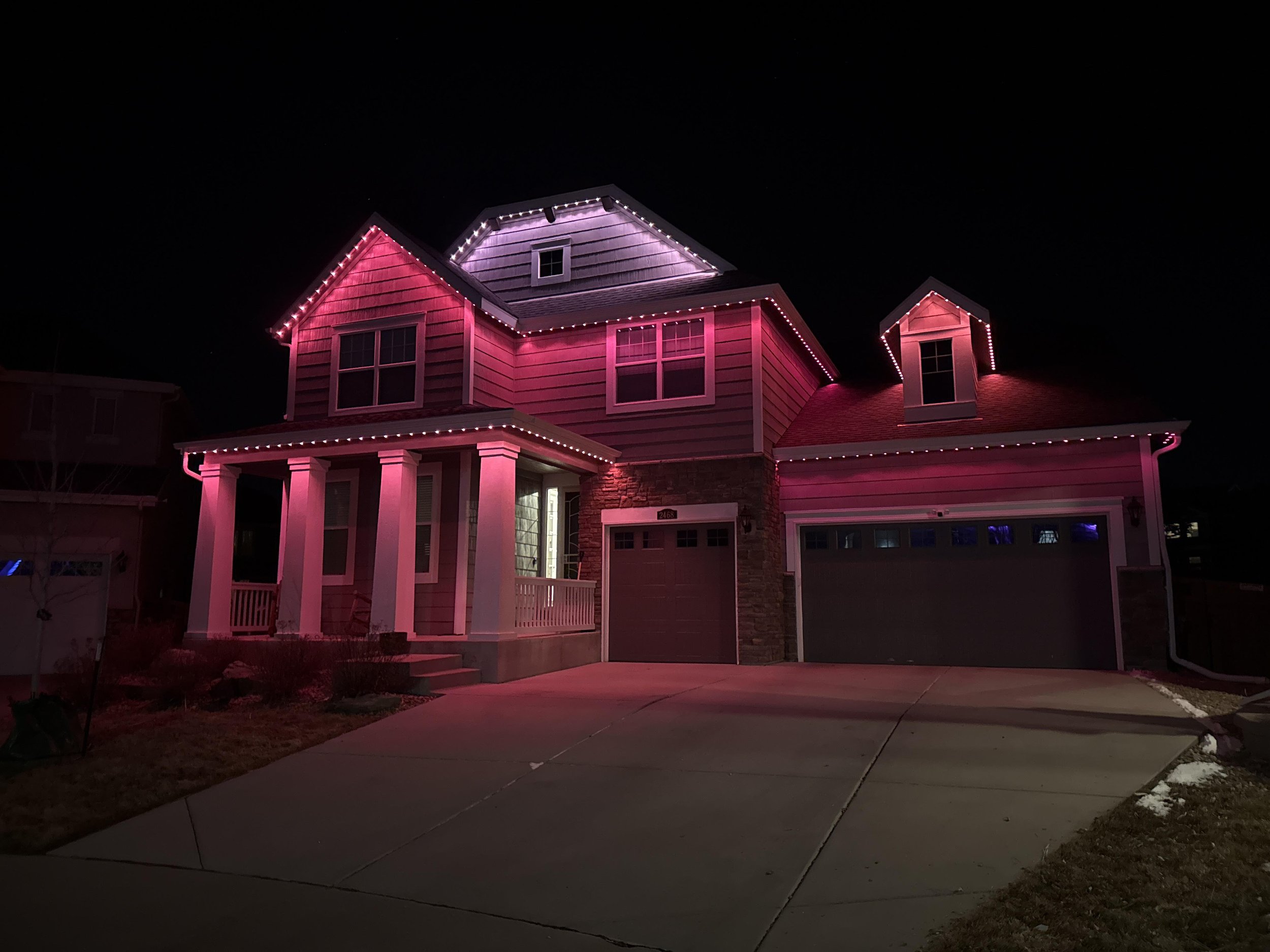 A two-story house decorated with pink and purple Christmas lights at night, with a sidewalk in front.