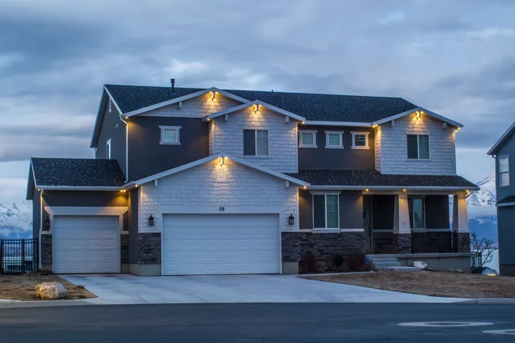 Modern two-story house with a double garage, exterior lighting, and a front porch, situated in a suburban neighborhood during dusk.