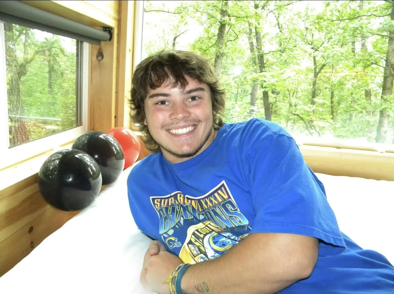 A young man with brown hair and a beard, smiling, leaning on a bed in a wooden cabin with windows showing a green forest. There are bowling balls on a ledge beside him.
