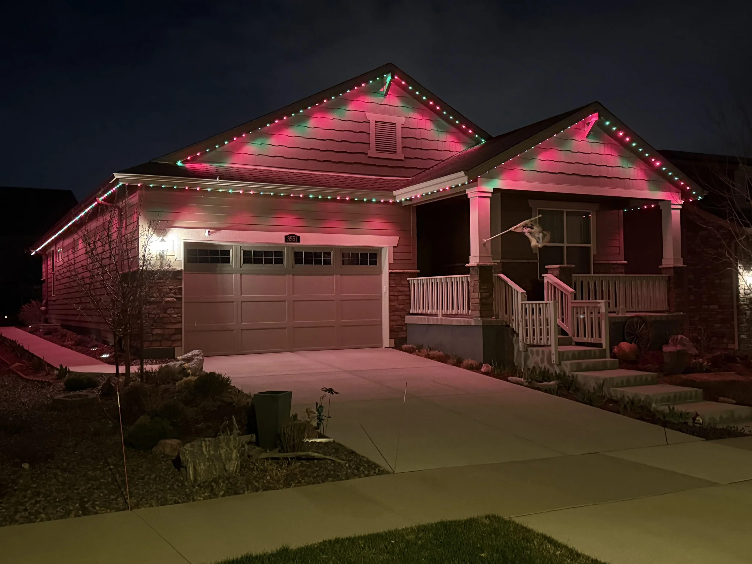 A house decorated with string lights along the roofline in red and green colors at night. The house has a garage door, a small porch with stairs, and a yard with a tree and potted plants.