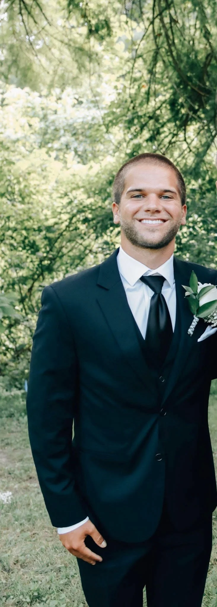 A man in a black suit, white dress shirt, and black tie smiling outdoors. He has short hair and a boutonniere on his lapel, with trees and greenery background.
