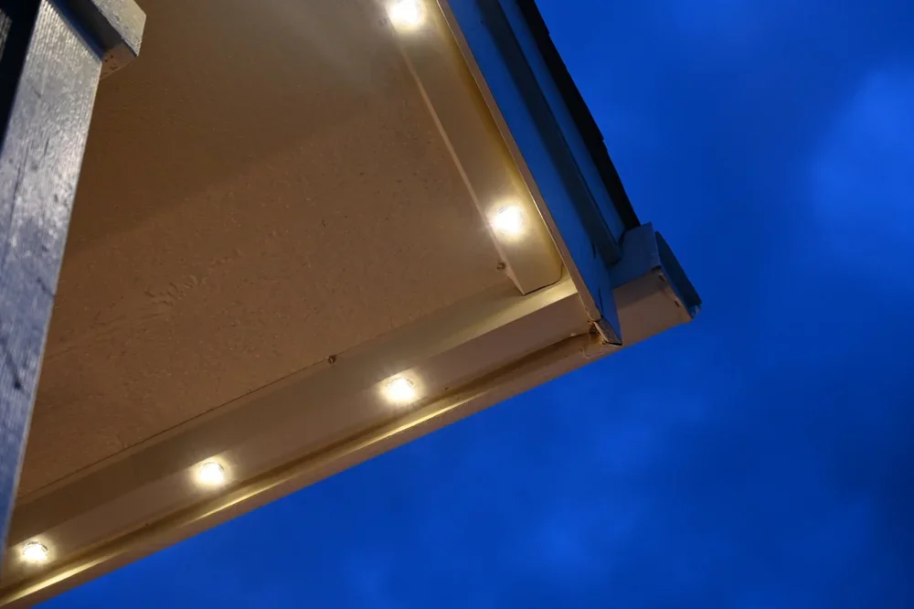 Close-up of a building's exterior corner at dusk with blue sky, featuring metal siding, a gutter, and a row of small round exterior lights.