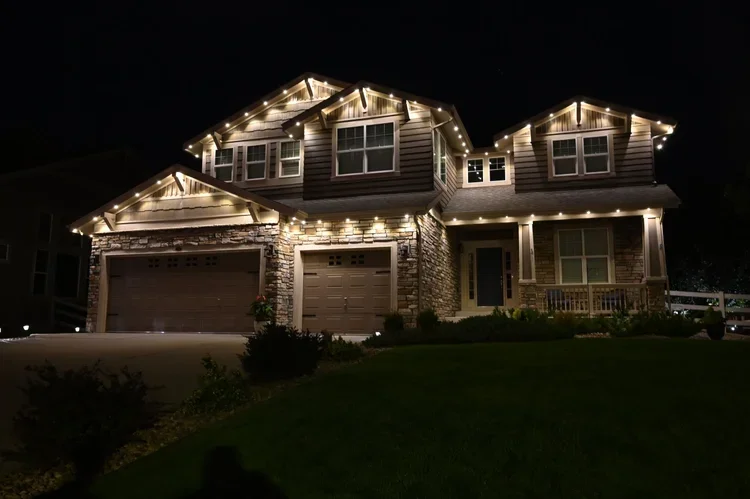 Two-story house at night with exterior lights and string lights along the roofline, stone and siding exterior, garage doors, front porch, and a well-maintained lawn.