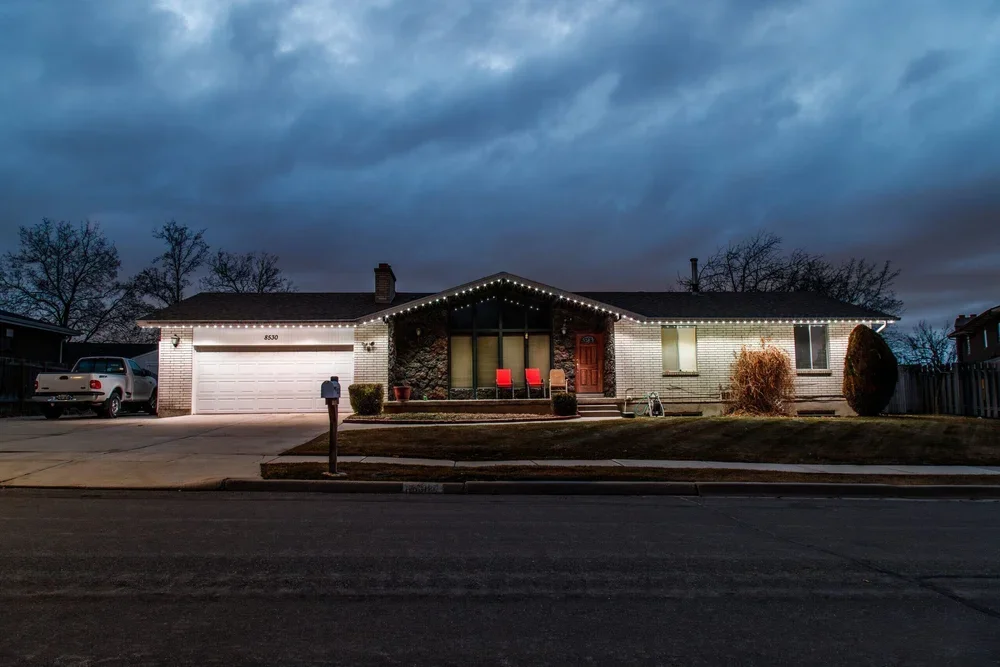Single-story house decorated with Christmas string lights on the roof, front porch, and garage. The house has a brick exterior, two red chairs on the porch, and is situated on a street with a parked pickup truck in the driveway. The sky is dark and cloudy.