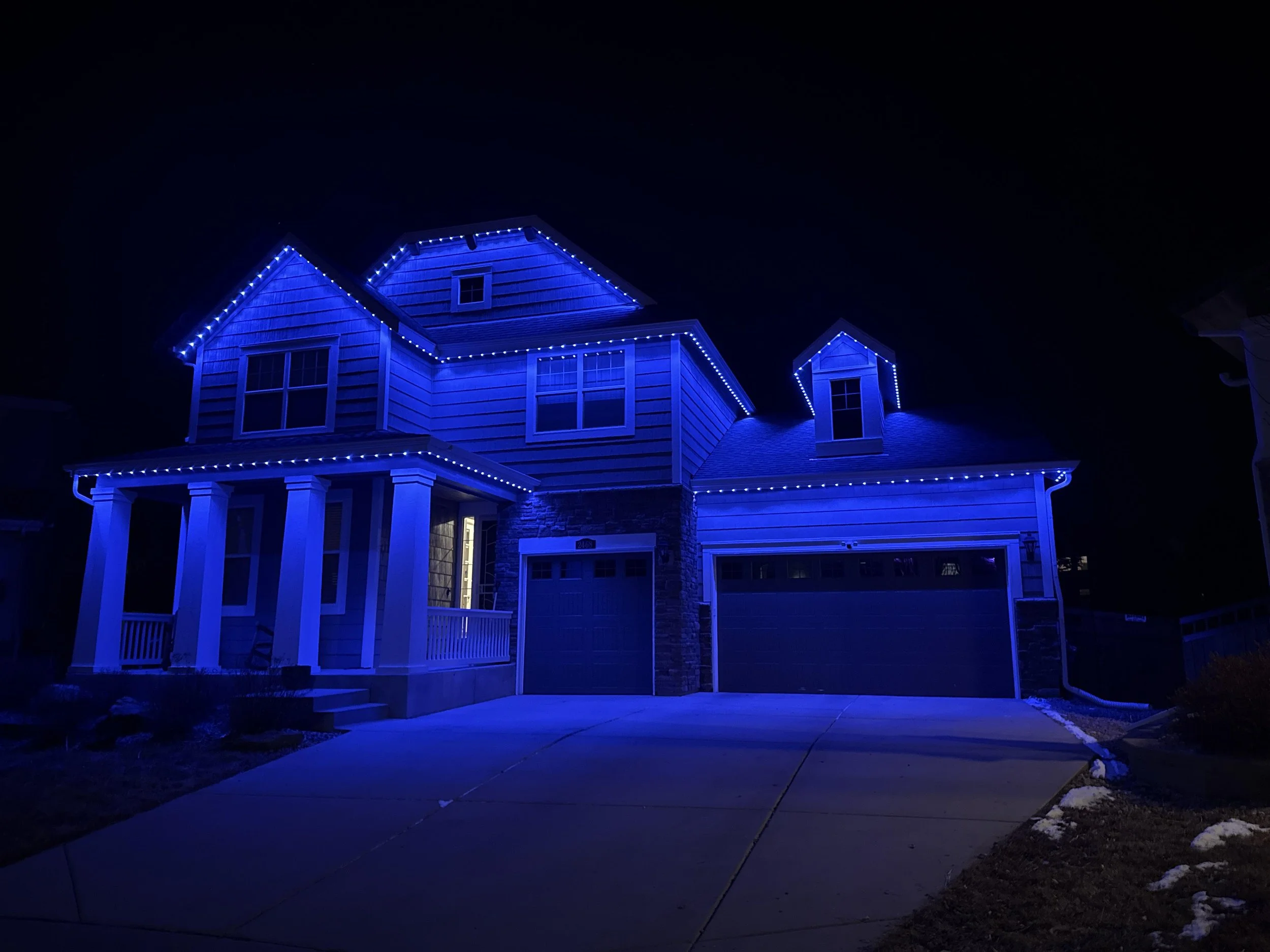A two-story house decorated with blue holiday lights along the roofline, windows, and porch at night. The house has white siding and a front porch with white columns, with a driveway and some snow on the ground.